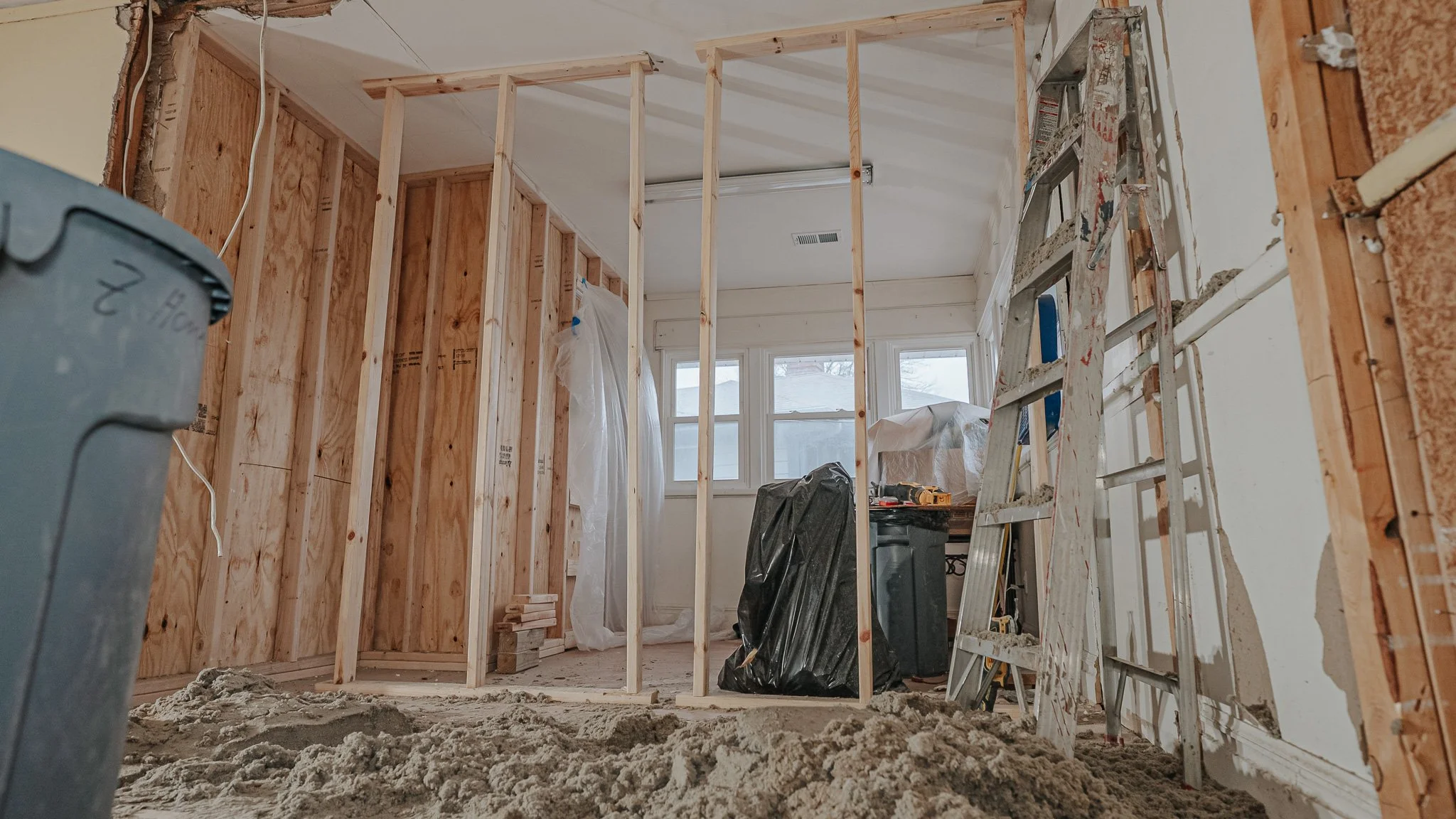Interior of a house under construction with exposed wooden framing, a ladder, a trash bag, and construction tools, with a window in the background.