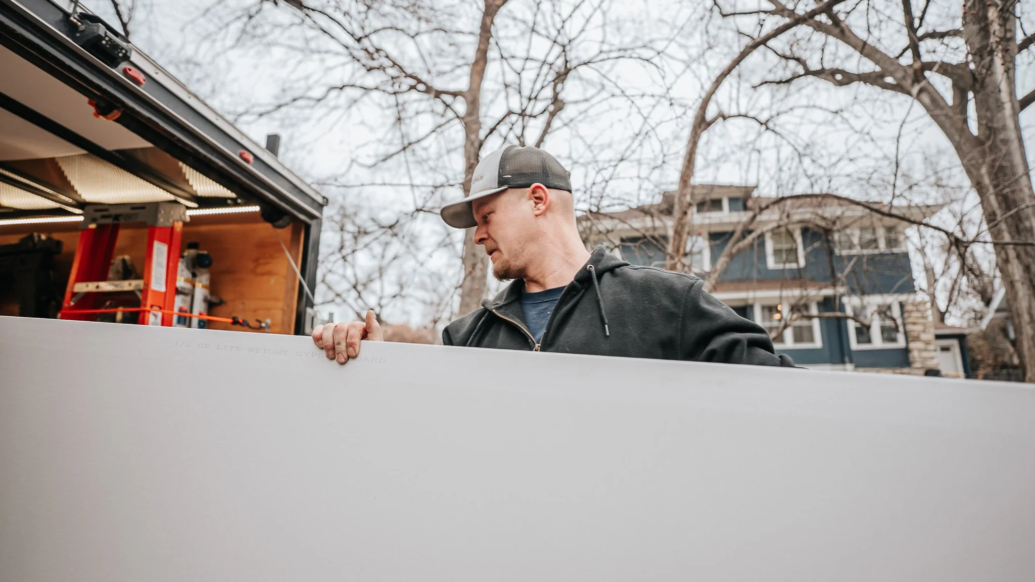 Man in black hoodie and cap loading a large white board or panel from a truck with open back, in a residential neighborhood with trees and houses in the background.