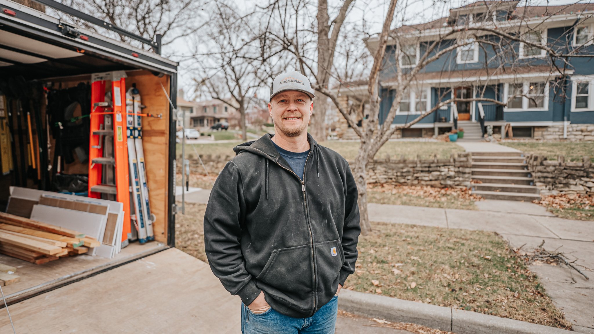 Smiling man standing outdoors in front of a house and a tree, wearing a black jacket and a gray cap, with a woodworking trailer filled with tools and wood behind him.