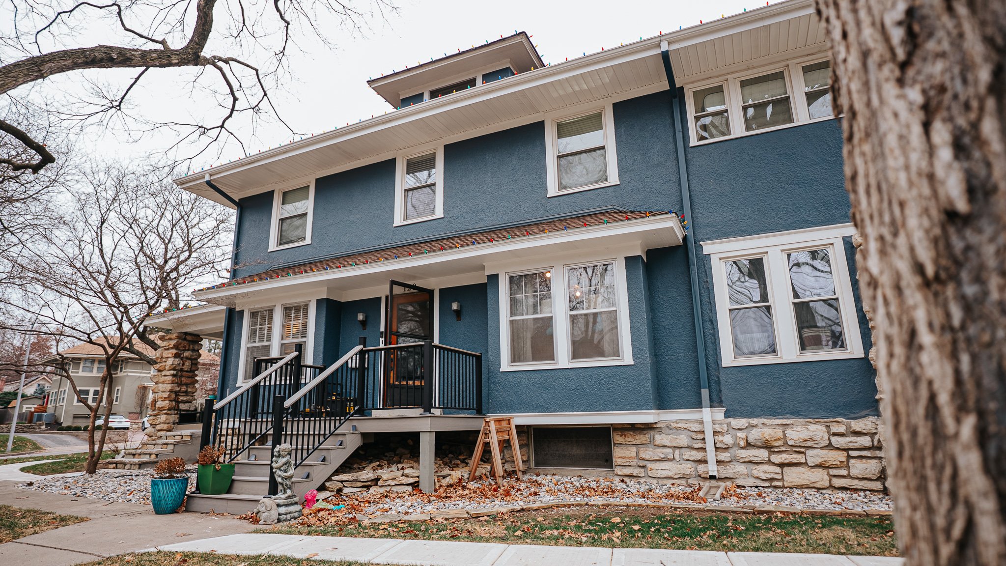 A blue multi-story house with white trim, decorated with holiday string lights along the roof. A front porch has black railing, a set of stairs, and is adorned with potted plants and a small statue. The yard has some rocks, a small tree, and a partially visible sidewalk.