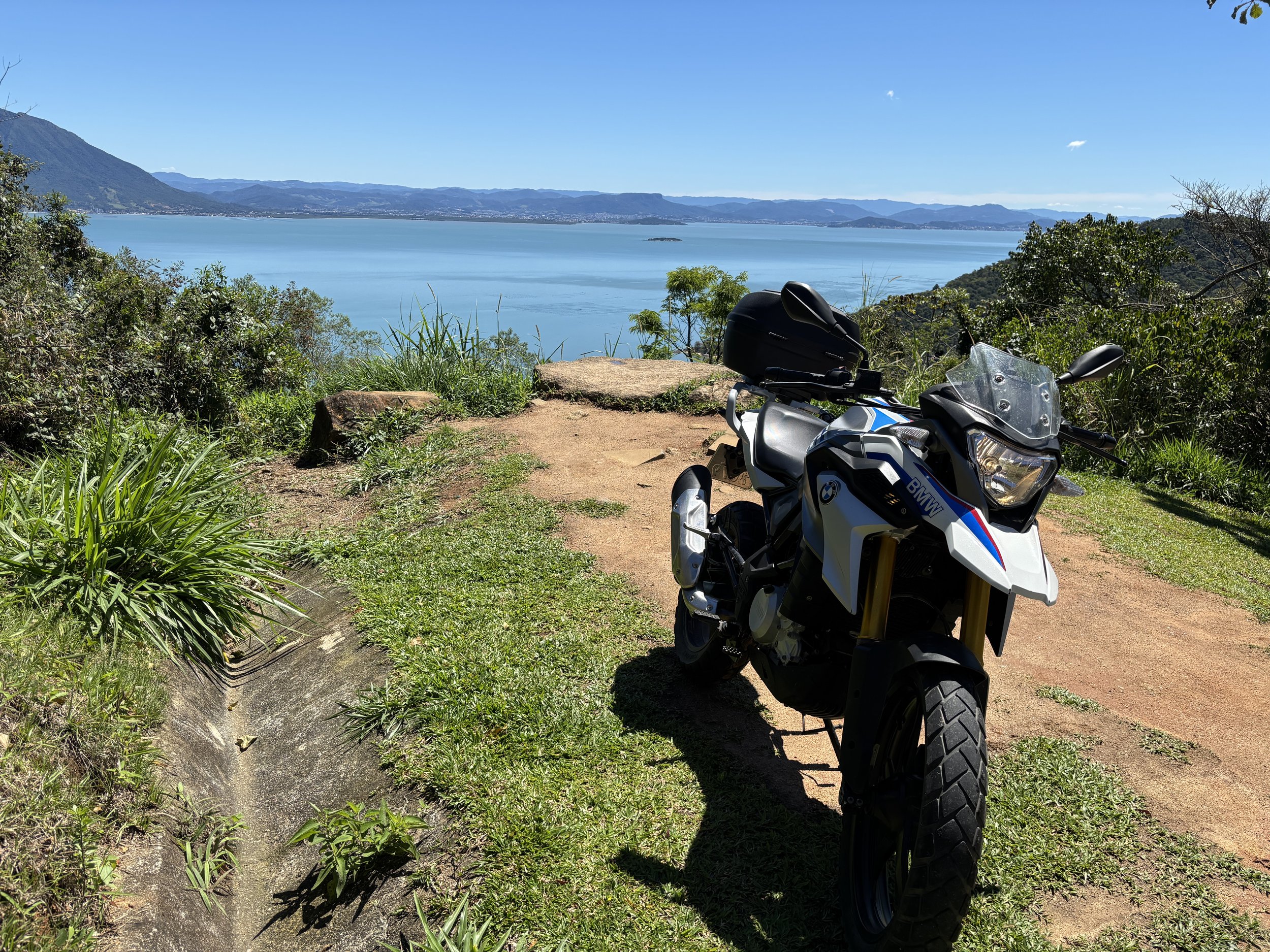A BMW rental motorcycle in Florianópolis and floripa parked on a dirt path near green foliage with a scenic view of the sea, mountains, and a clear blue sky in the background. alugar um moto em floripa