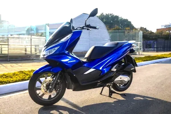 Scooter azul disponível para aluguel em Florianópolis, estacionada em área urbana com vegetação ao fundo.