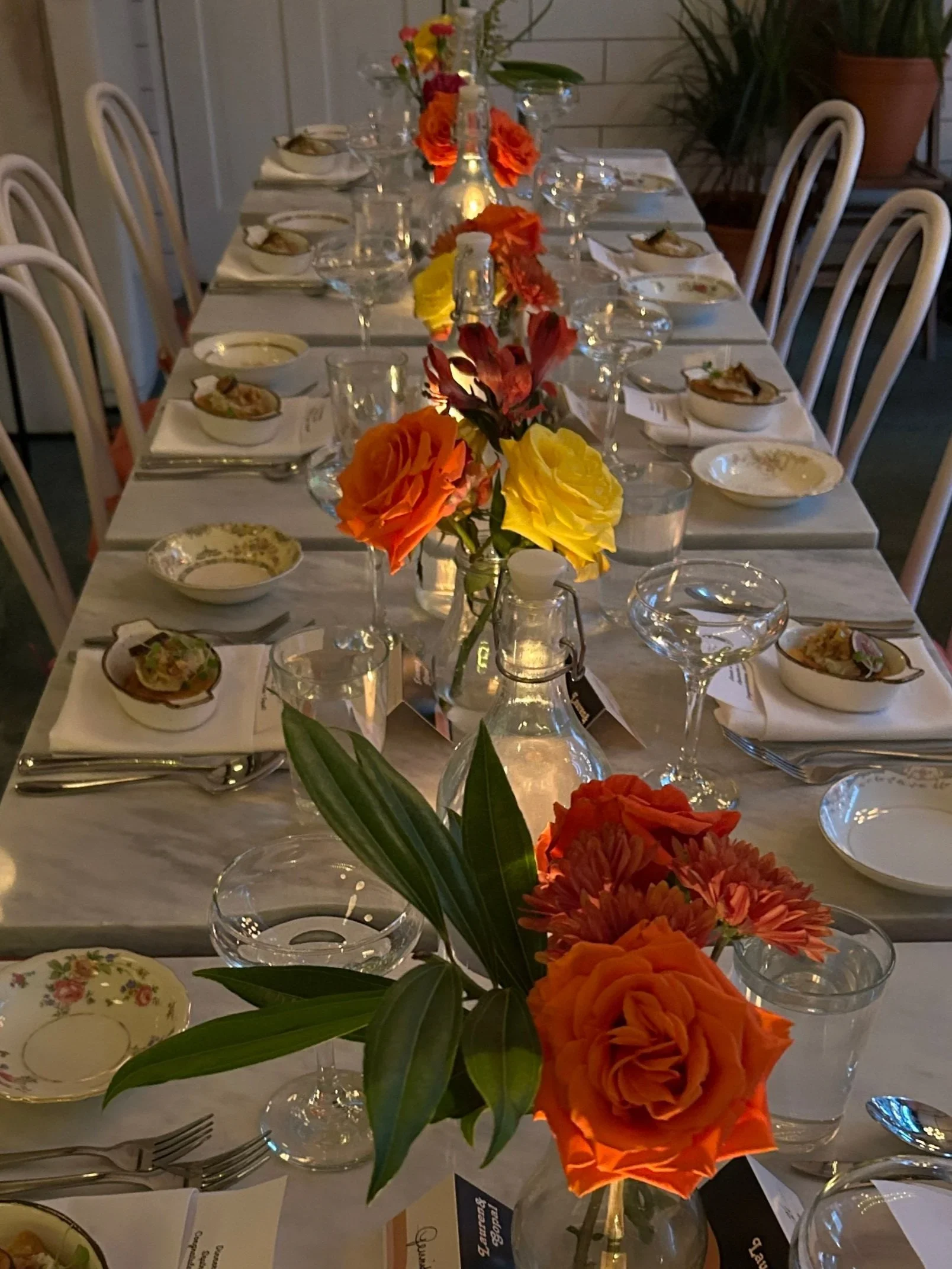 Long dining table set for a special occasion with floral centerpieces in orange, yellow, and red, surrounded by elegant place settings, candles, and glassware.