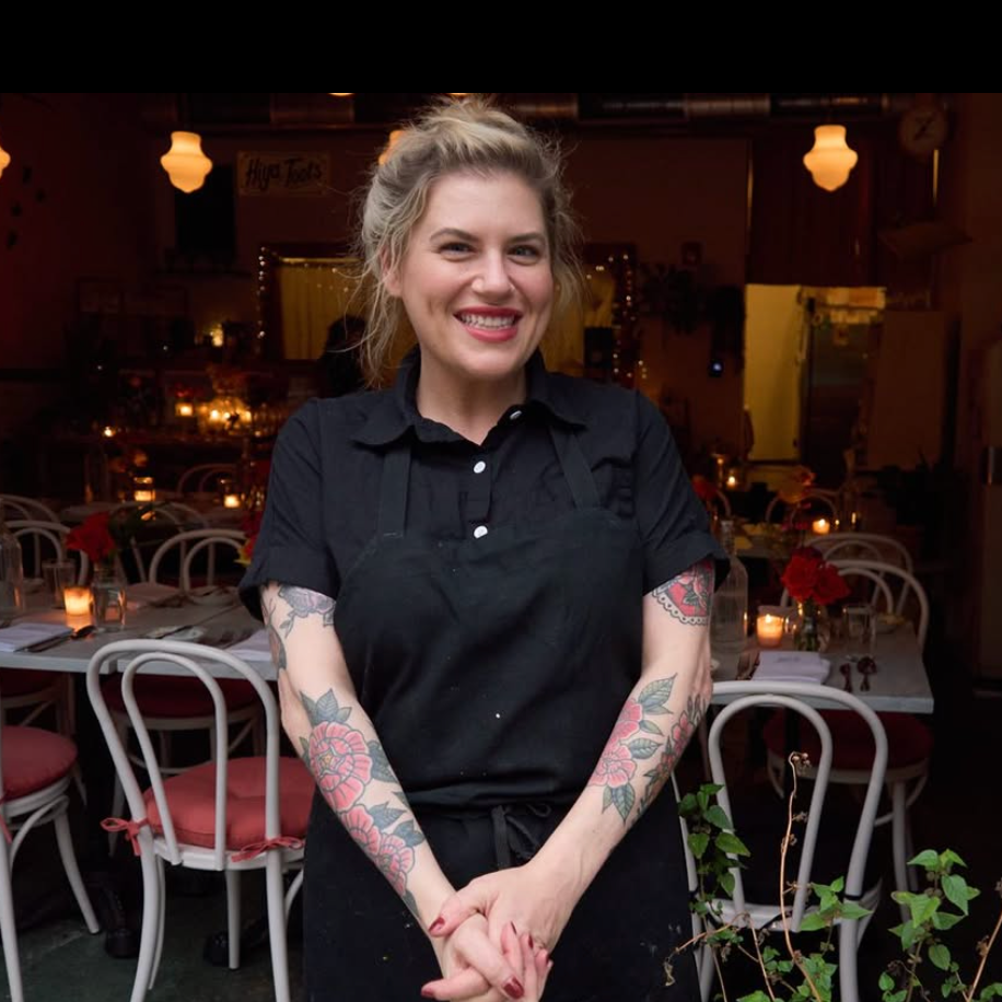 A smiling woman with tattoos on her arms, wearing a black shirt and apron, standing in a warmly lit restaurant with decorated tables and candles.
