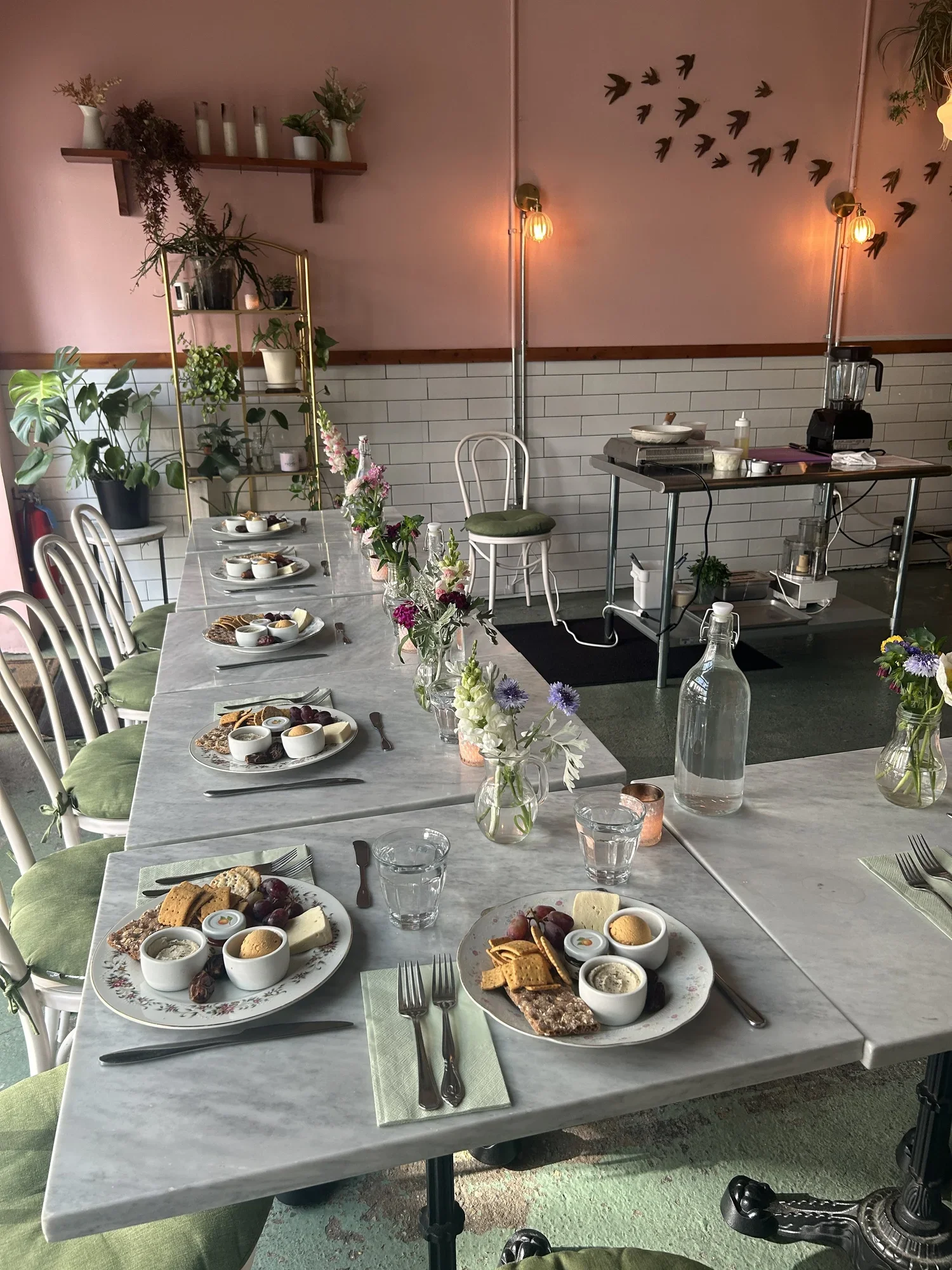 A decorated dining table set for a meal with plates of cheese, crackers, grapes, and small bowls of dips or spreads, surrounded by chairs with green cushions, with flowers in vases and a pink wall with wall art of flying birds in the background.