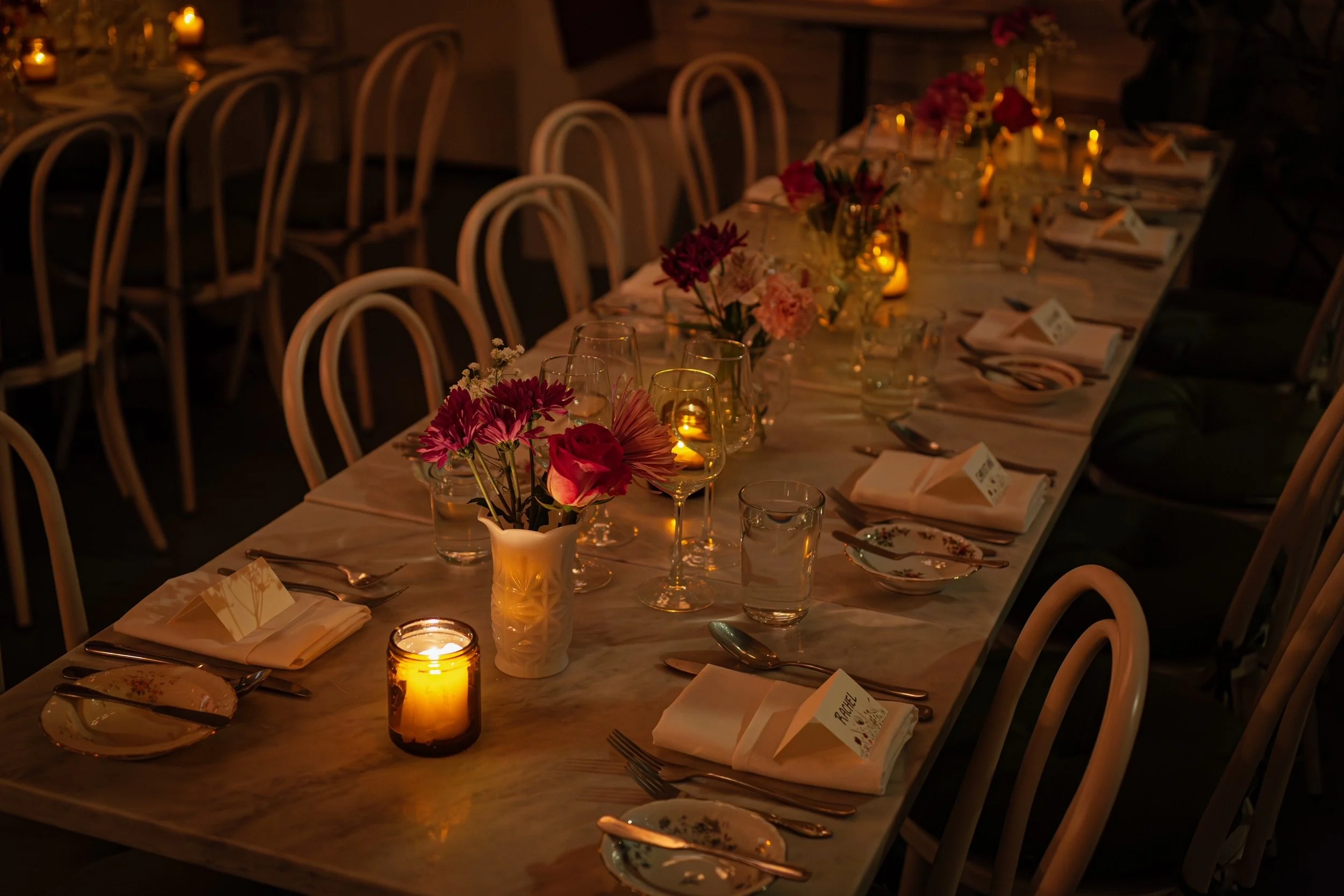 A long dining table set for a formal dinner with floral centerpieces, candles, glassware, and folded napkins, in a dimly lit ambiance.