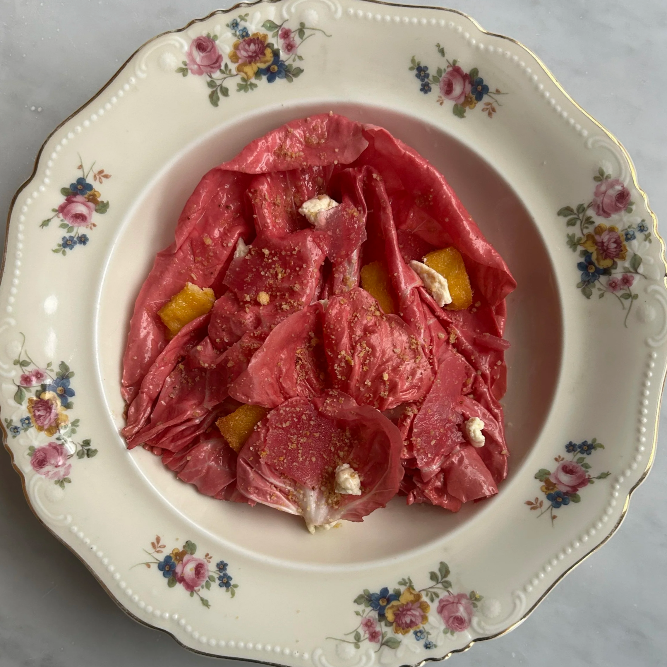 A vintage floral plate with pink, purple, and yellow flowers along the border, holding a dessert with pink or red flower petals, yellow cubes, white crumbles, and a sprinkle of gold powder.
