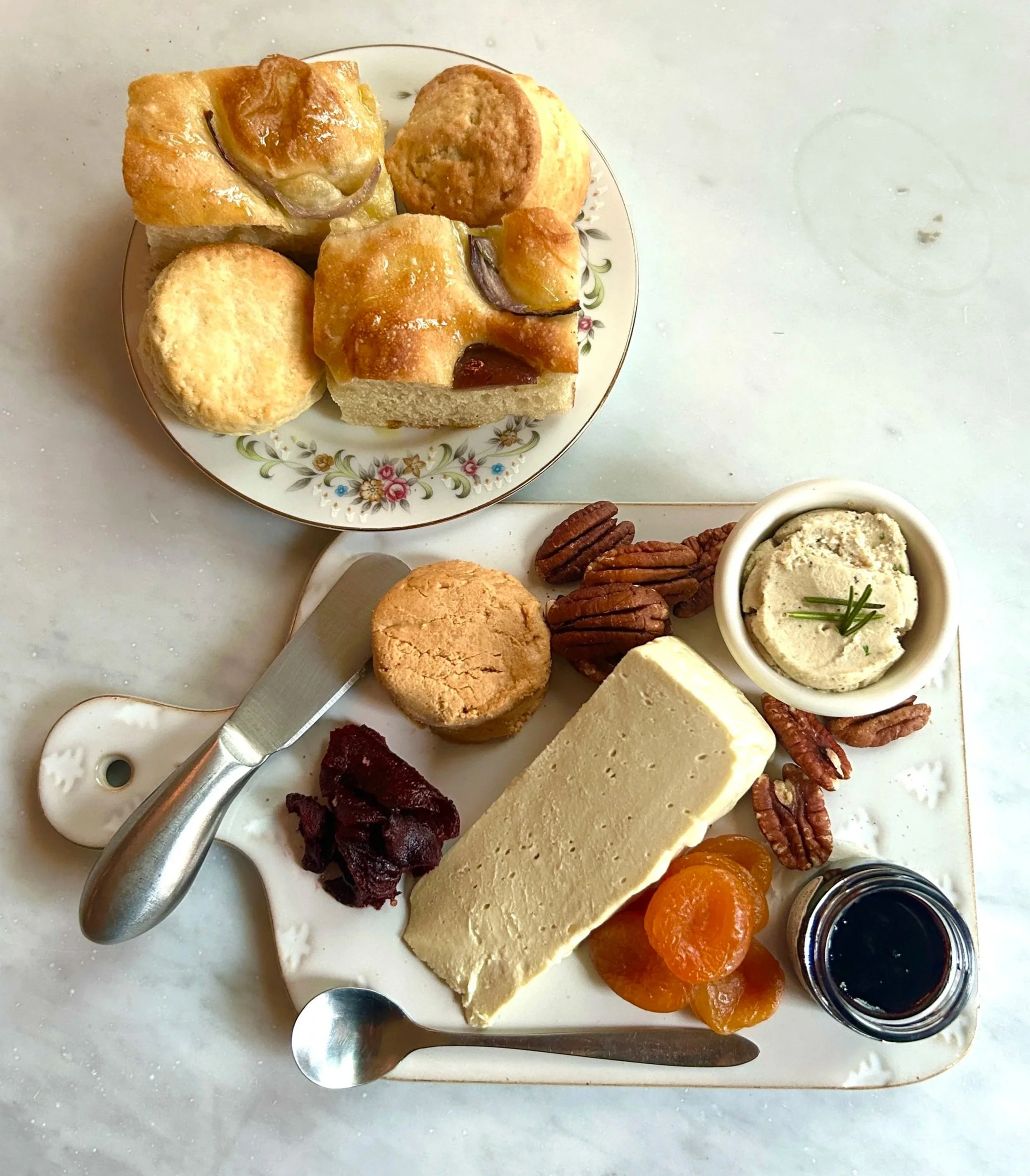 A cheese platter with assorted cheeses, pecans, dried apricots, fig jam, and a small bowl of herb spread, alongside a serving of scones on a floral plate.