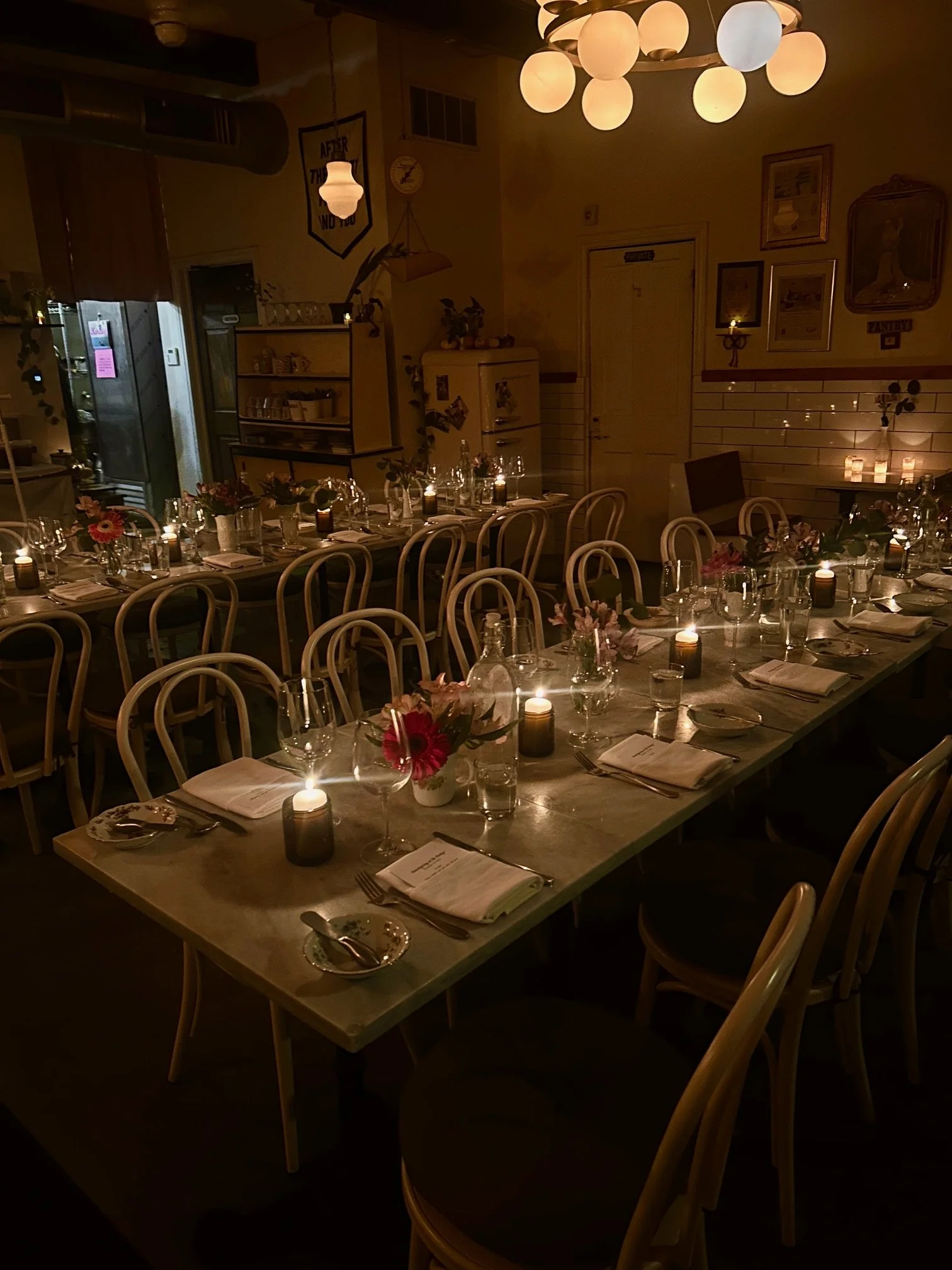 A dimly lit dining room with a long table set for a dinner, decorated with candles and floral centerpieces, surrounded by curved-back chairs, with warm lighting and vintage decor on the walls.