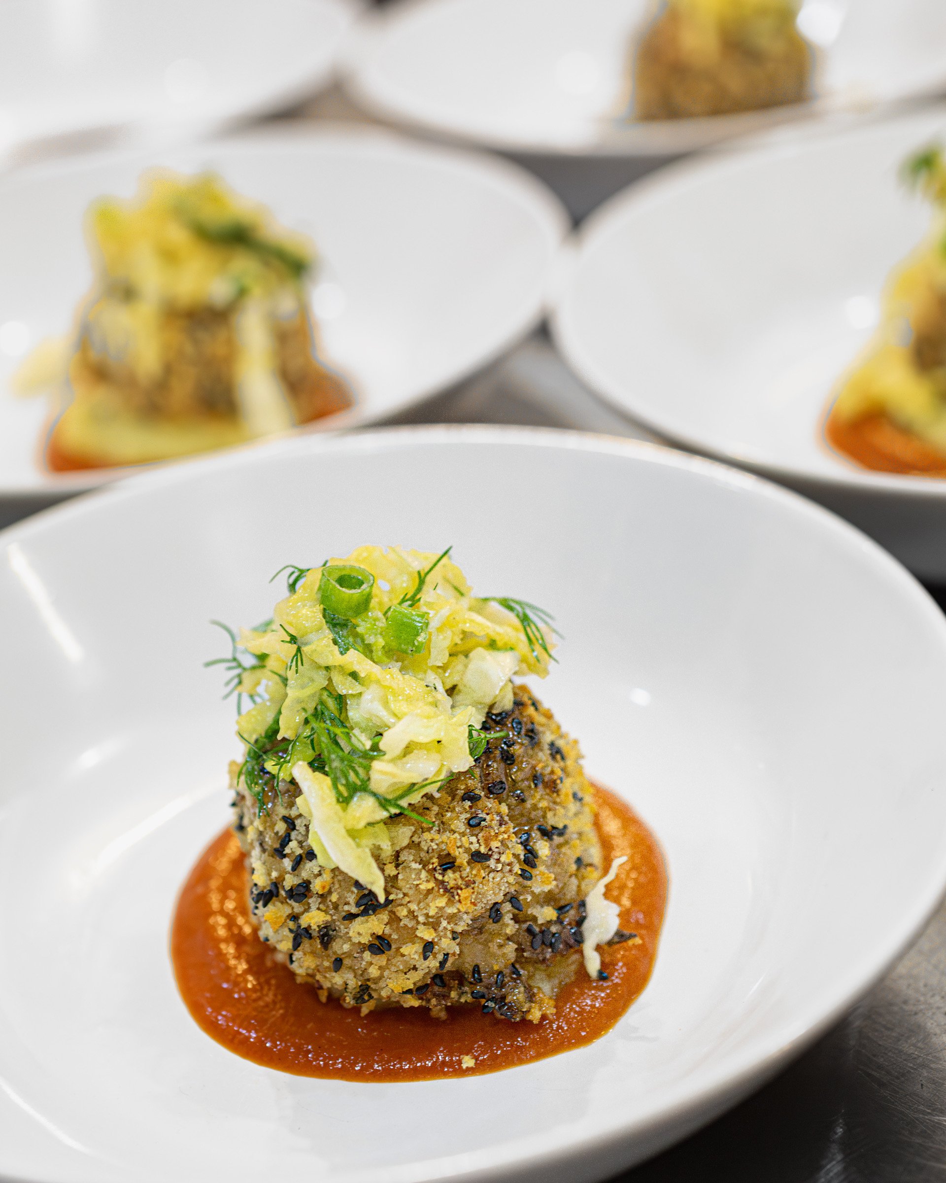 A plated gourmet appetizer with a breadcrumb-coated ball topped with chopped vegetables and green herbs, served on a red sauce in a white dish.