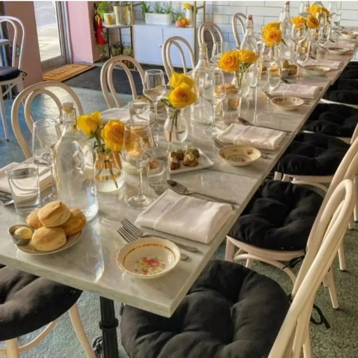 Dining table set for a meal with yellow roses in glass vases, water bottles, wine glasses, plates, napkins, and black chairs with cushions.