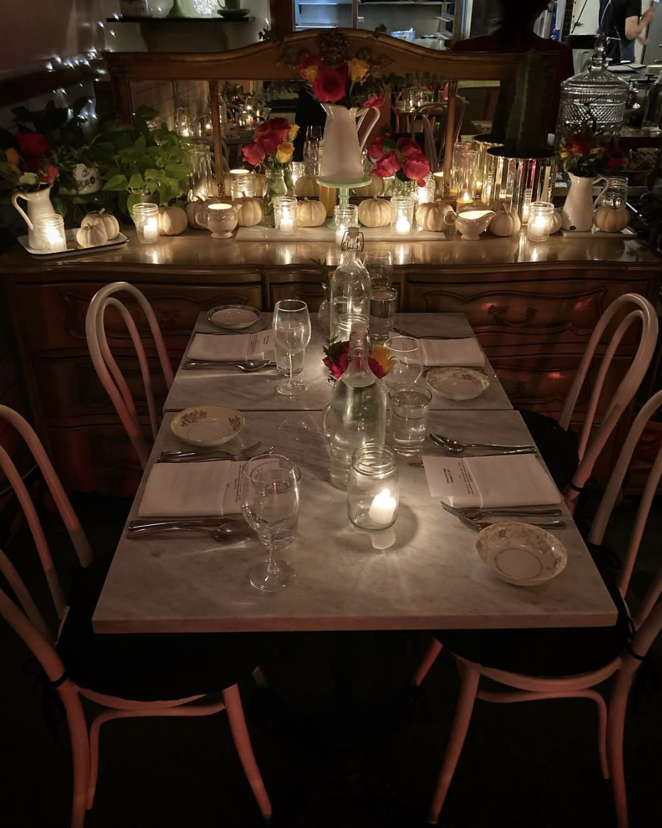 A decorated dining table with a marble top, set for a meal, illuminated by candles. The table features glass bottles as centerpieces, with flowers in shades of red, pink, and yellow. In the background, there is a wooden sideboard adorned with white pumpkins, candles, and floral arrangements, reflecting a cozy, warm ambiance.