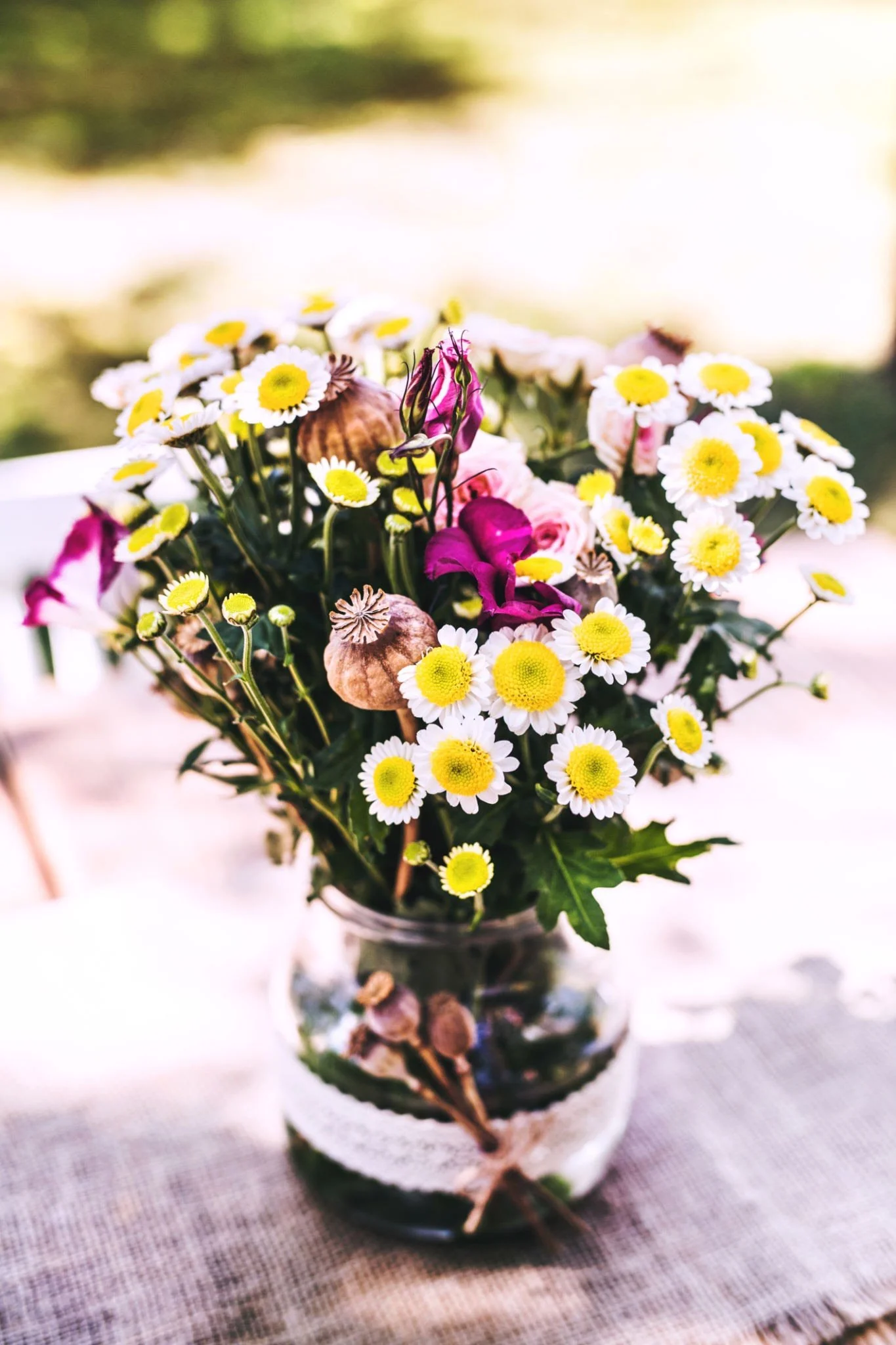 A glass vase filled with a bouquet of white daisies with yellow centers, pink flowers, and dried seed pods placed on a tablecloth outdoors.