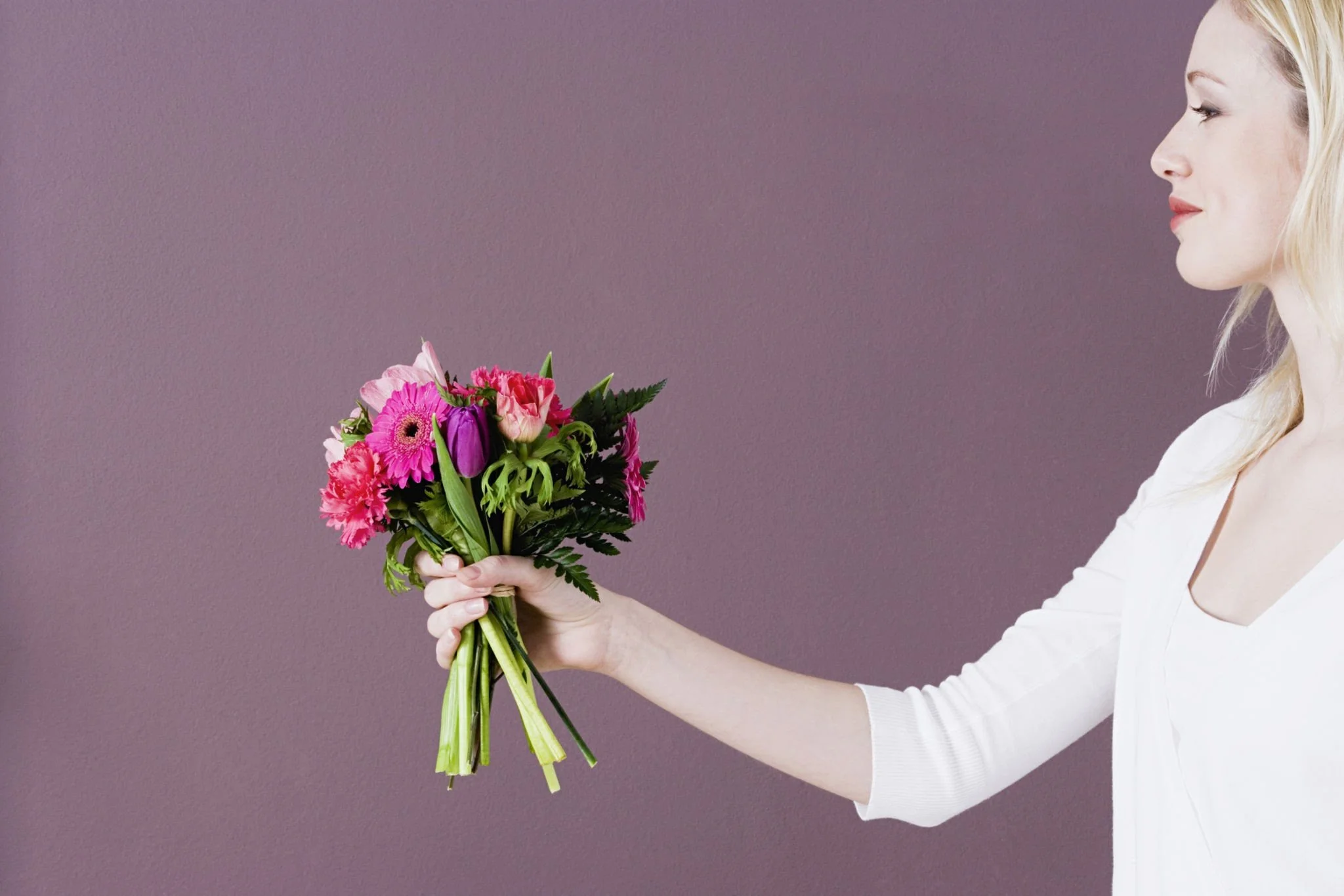 A woman with blonde hair, dressed in white, holding a colorful bouquet of flowers against a purple background.