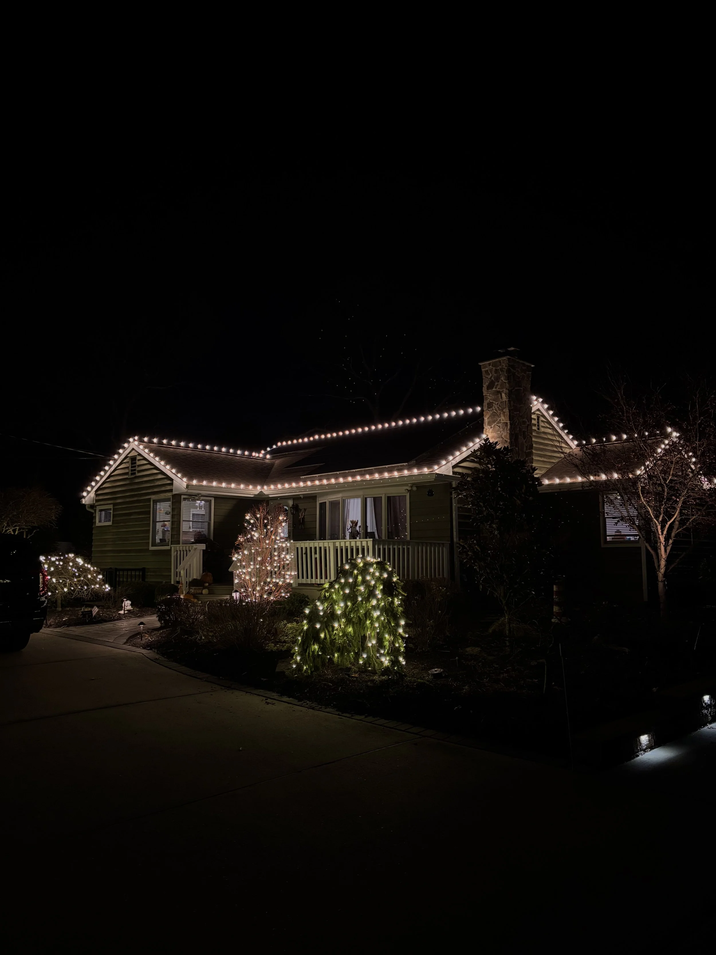 A house decorated with Christmas lights at night, featuring a small porch with a railing, illuminated bushes, and string lights outlining the roof.