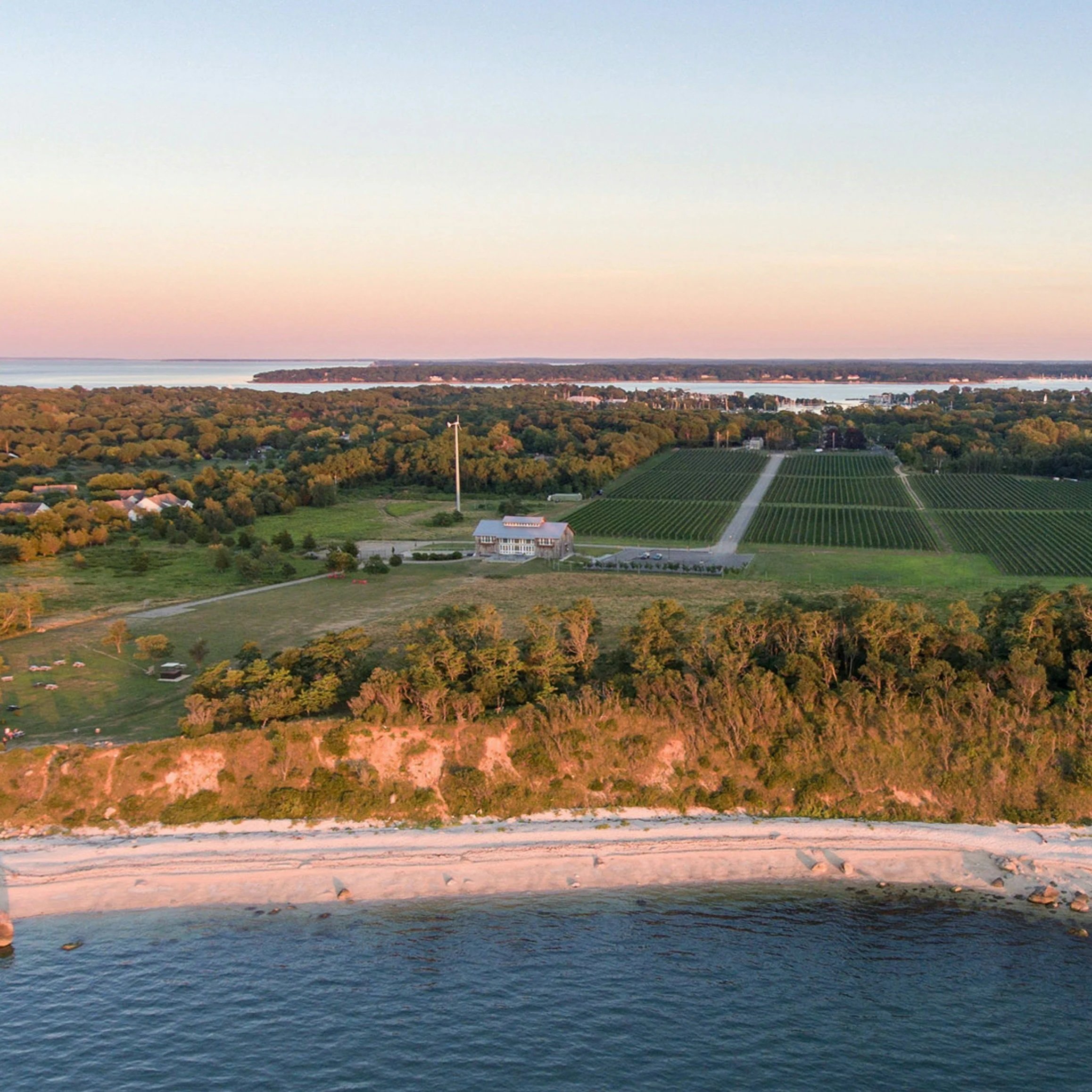 Aerial view of a coastal landscape during sunset with a beach, trees, fields, a building, wind turbines, and a body of water in the background.