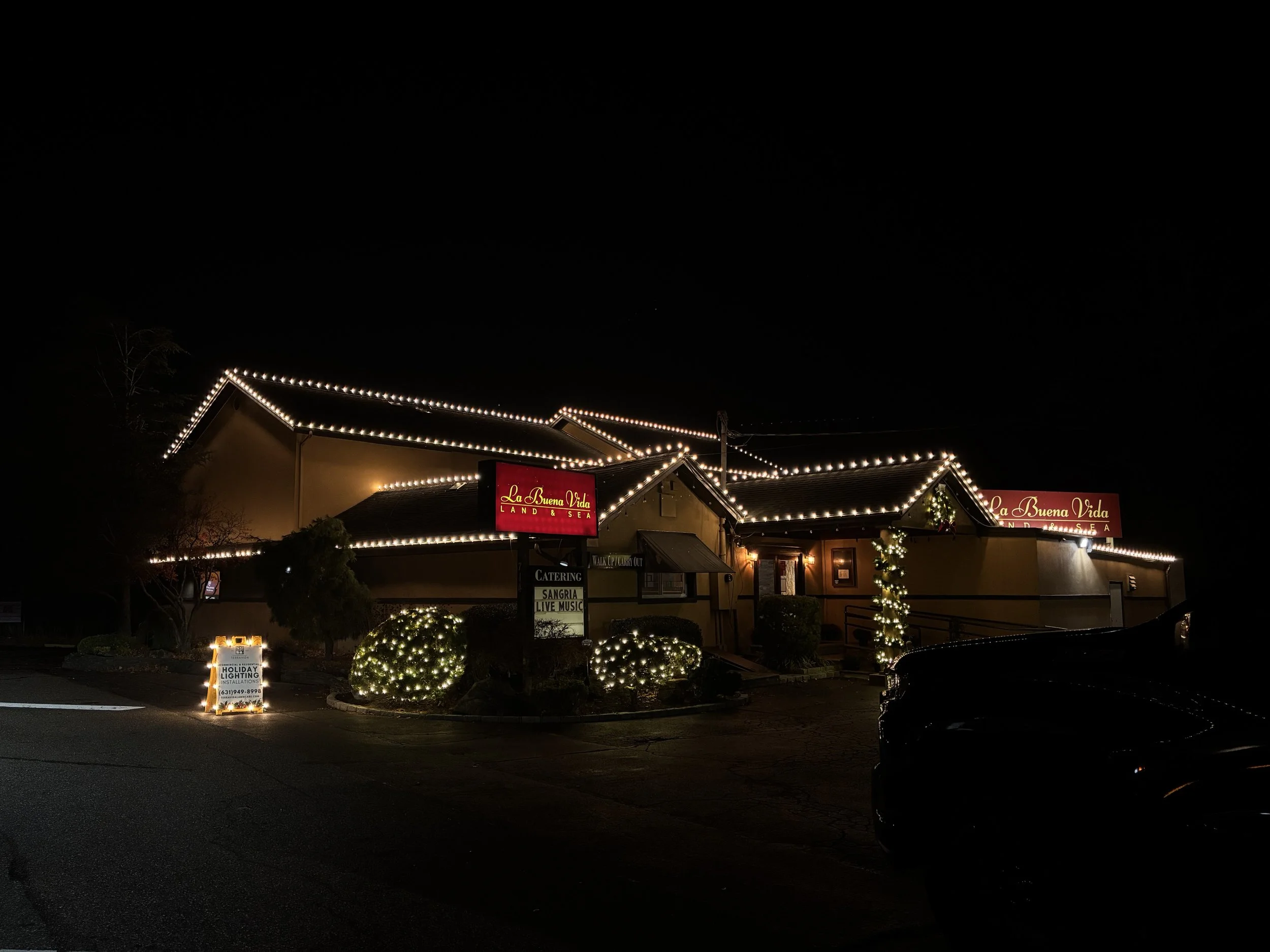 A restaurant decorated with string lights along the roofline at night, with bushes wrapped in lights, a sign that reads 'La Buena Vida,' and a small illuminated sign in the parking lot.