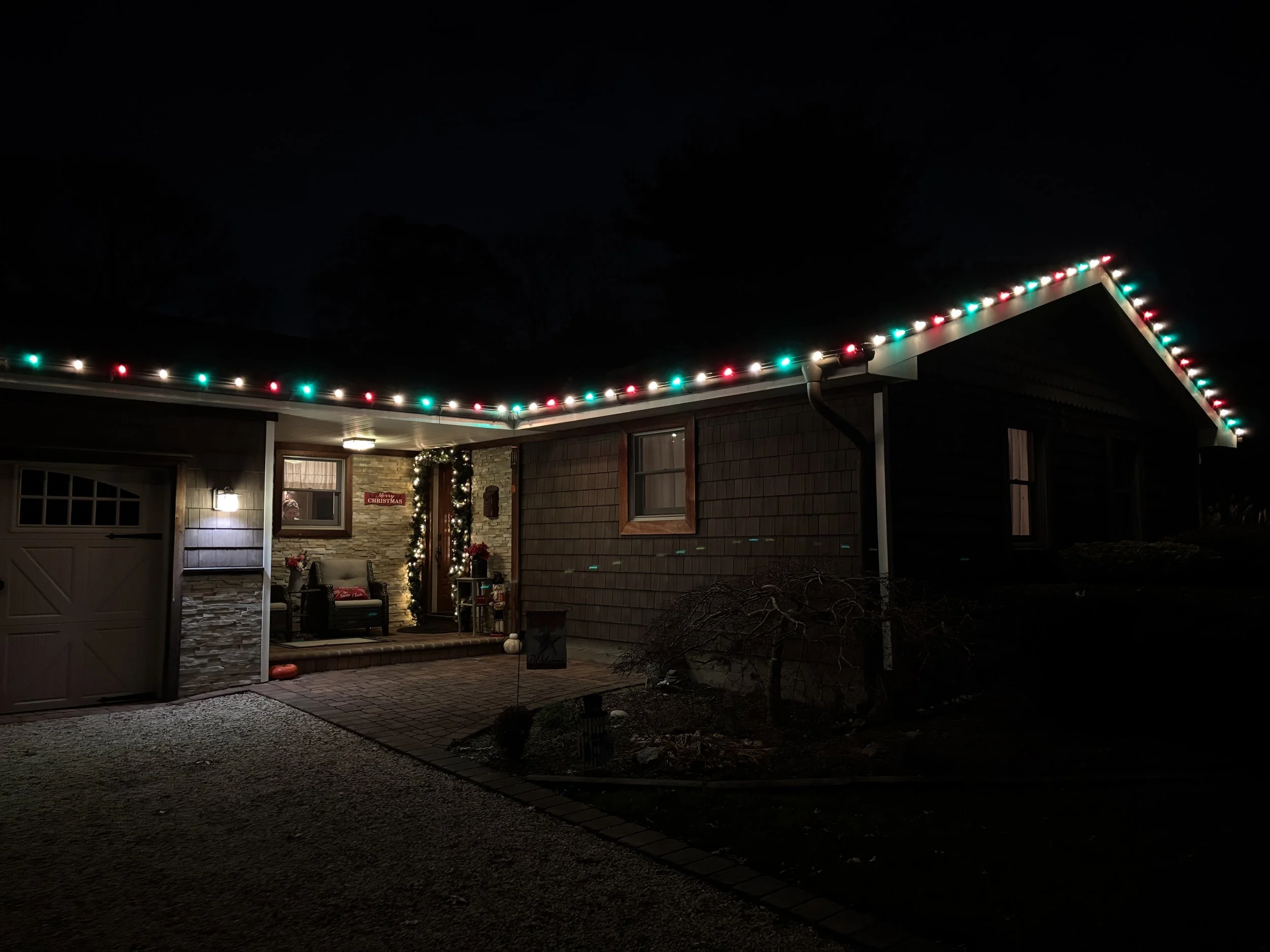 House decorated with multicolored Christmas lights along the roofline at night, with a front porch illuminated and decorated with Christmas wreaths and ornaments.