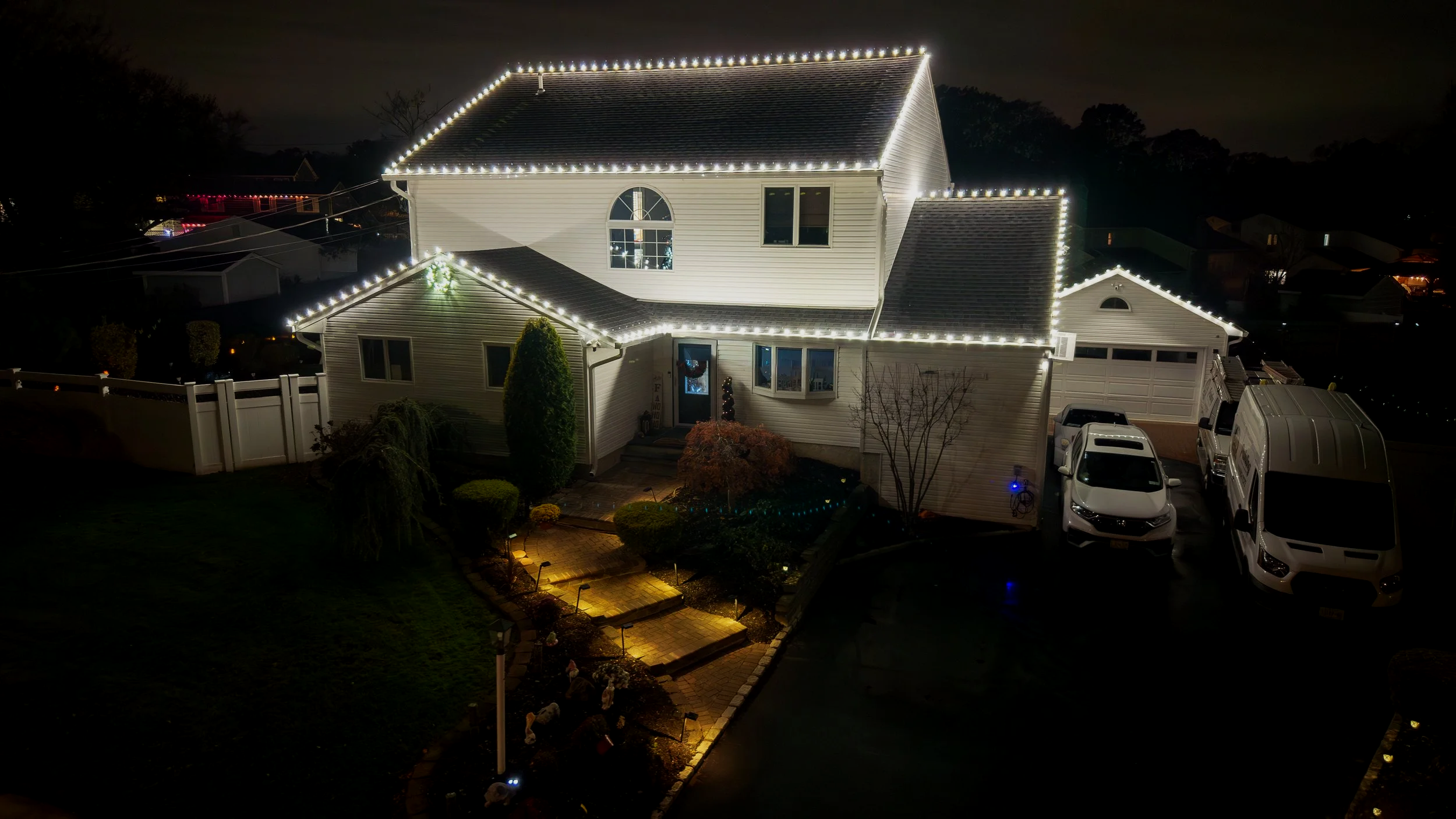 A two-story house decorated with white Christmas lights along the roofline and eaves at night. The house has a front yard with a walkway and garden, and a driveway next to parked cars, including a white van. The yard features bushes, trees, and garden lights.