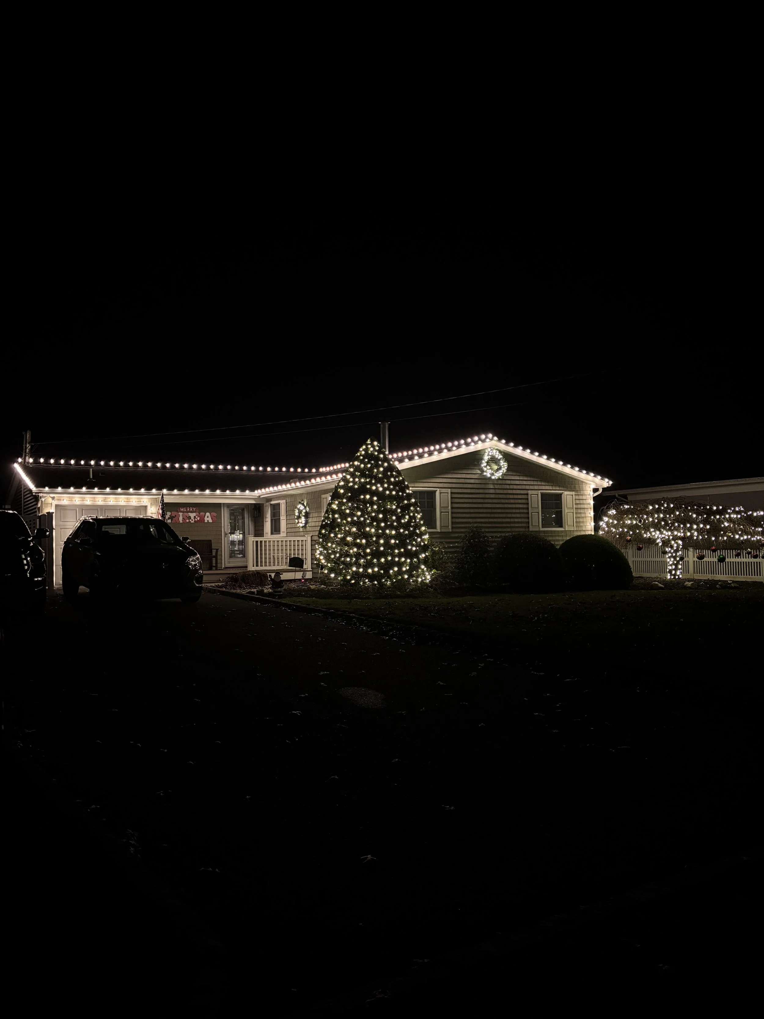 House decorated with Christmas lights and wreaths, a lit Christmas tree in the yard, and cars parked in the driveway at night.