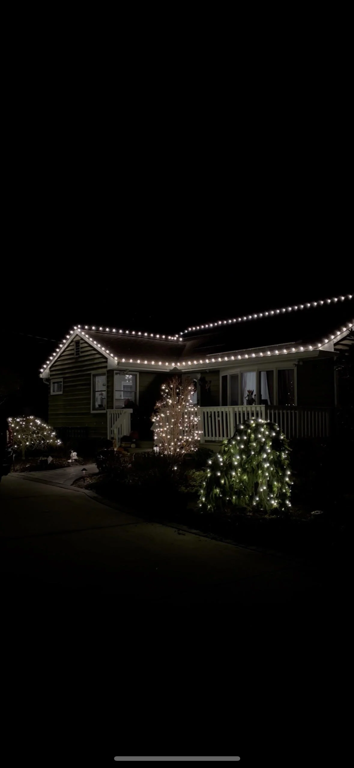 A house decorated with Christmas string lights along the roofline and around the bushes in the front yard at night.