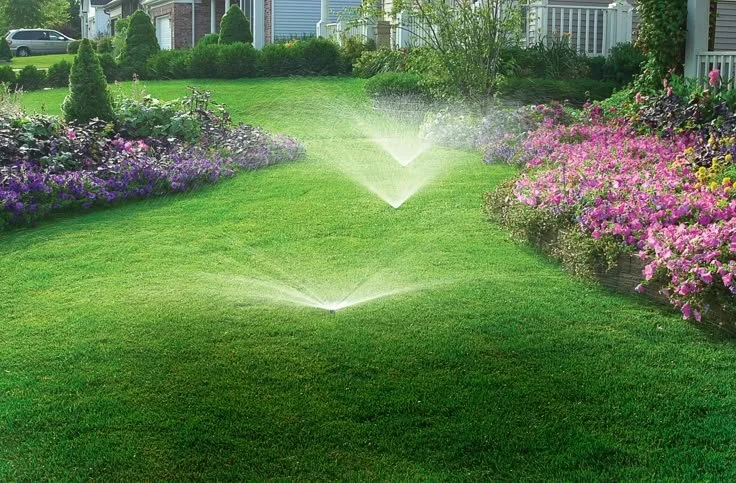 A lush, green front yard with sprinklers watering the grass, surrounded by colorful flower beds and a house in the background.