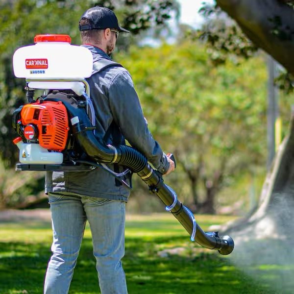 A man is using a backpack leaf blower in a park or backyard with green trees and grass.