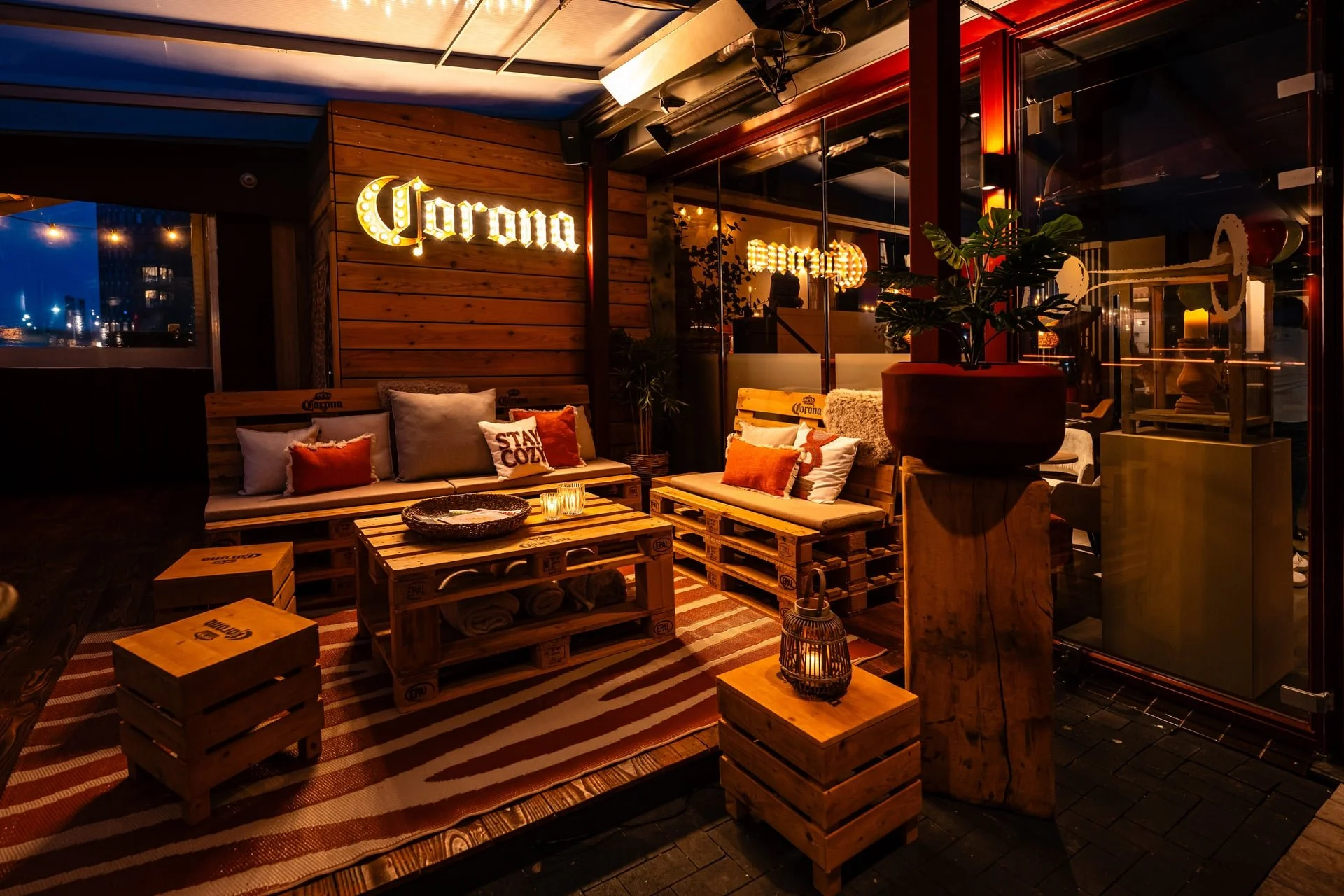 A cozy outdoor seating area of a bar or restaurant featuring wooden pallet furniture, cushions, a striped rug, decorative candles, and a large potted plant. The area is lit with warm lighting and a neon sign that reads 'Corona' in the background.