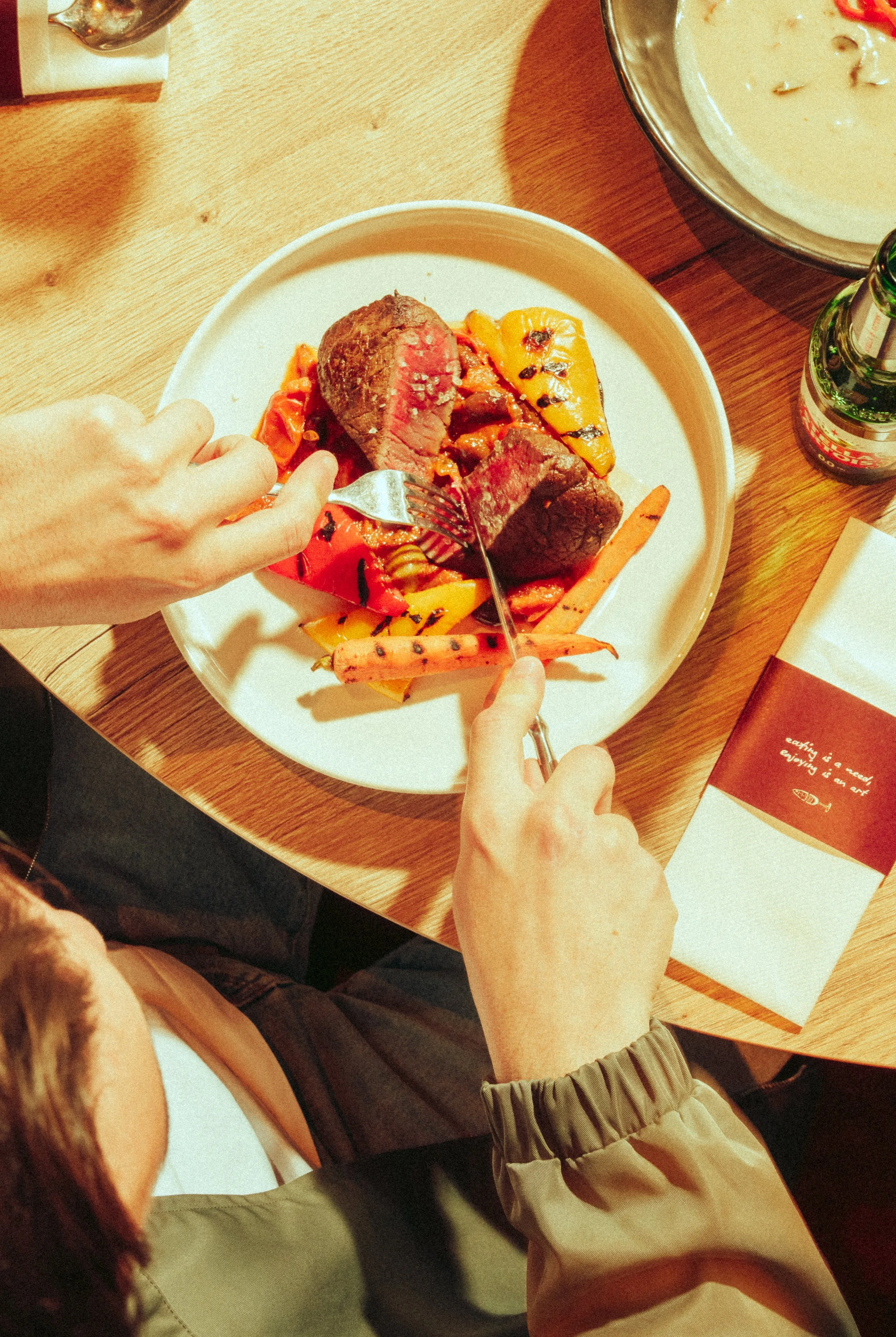 Person cutting medium-rare beef steak with grilled vegetables on a white plate at a wooden table.