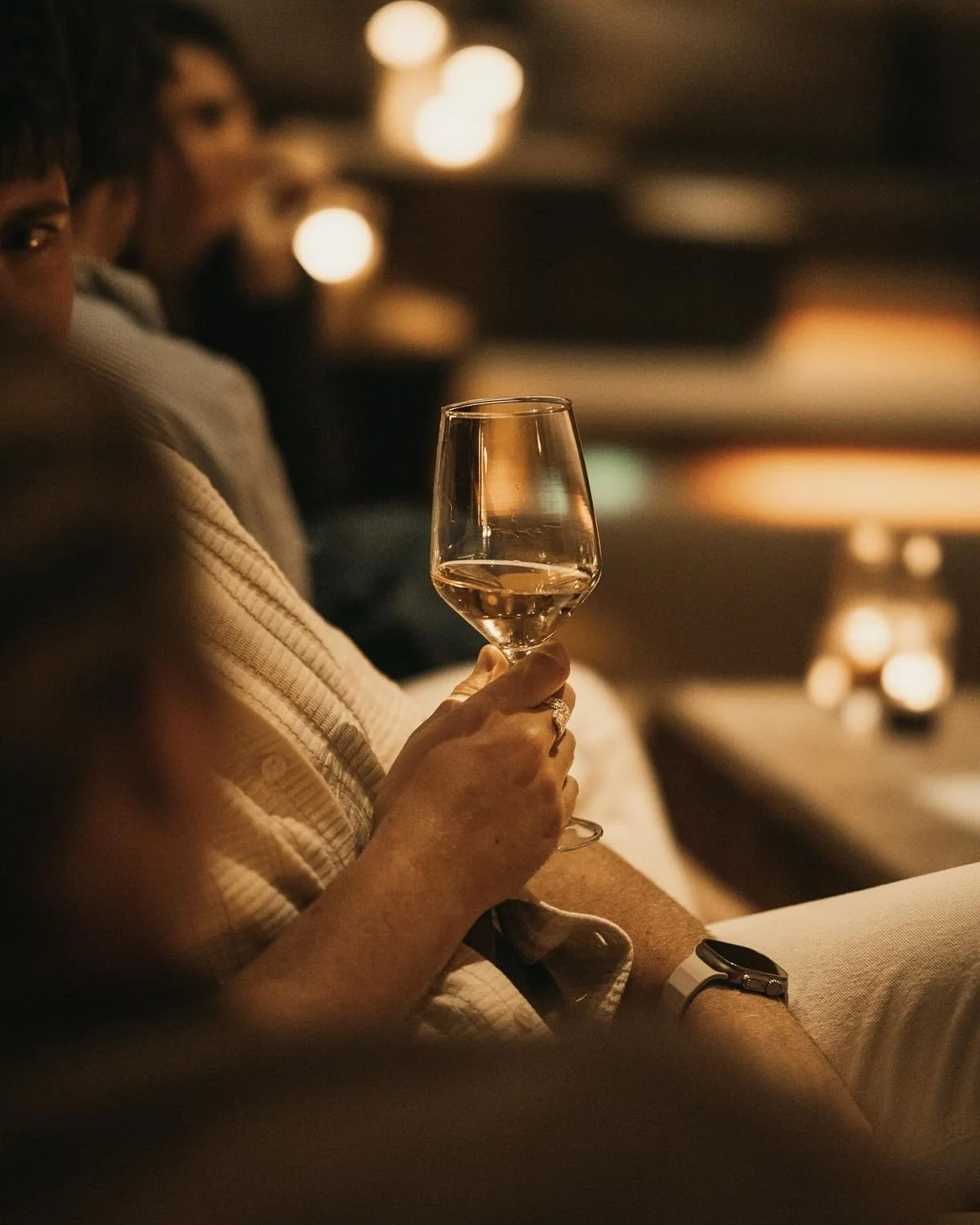 Person holding a wine glass in a dimly lit setting, with other people and warm lighting in the background.
