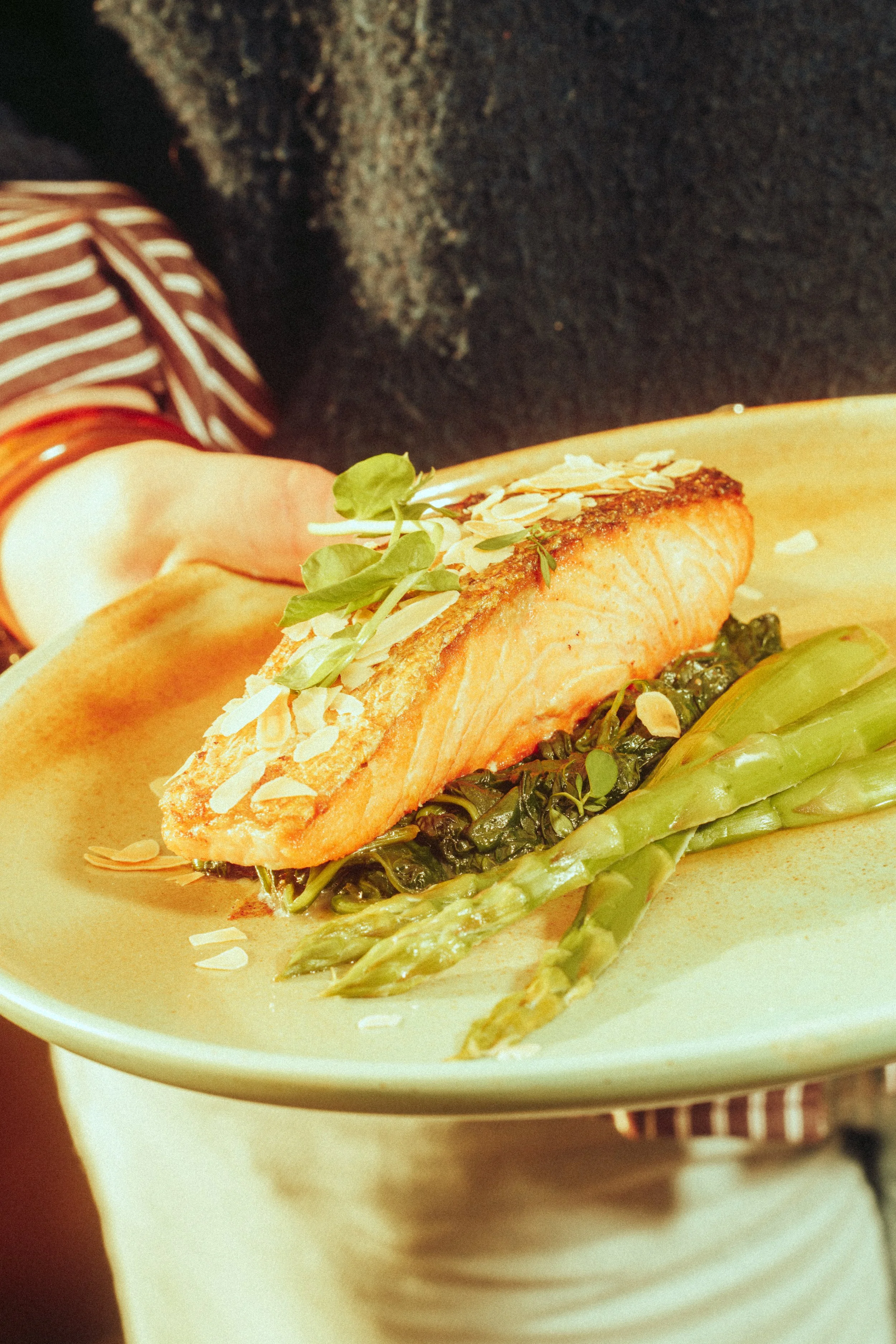A plated dish with a seared piece of salmon, garnished with sliced almonds and microgreens, served on top of sautéed greens with a side of steamed asparagus.