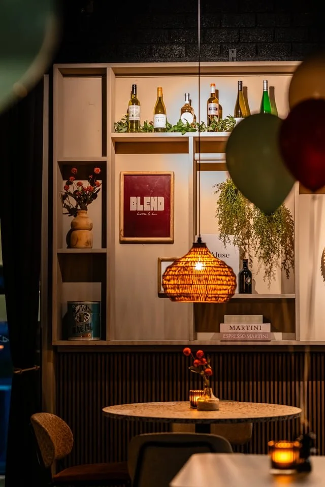 Cozy dining area with a round table, a rattan pendant light, and decorative balloons. Shelves with wine bottles, a framed art piece, a vase with red flowers, and greenery.