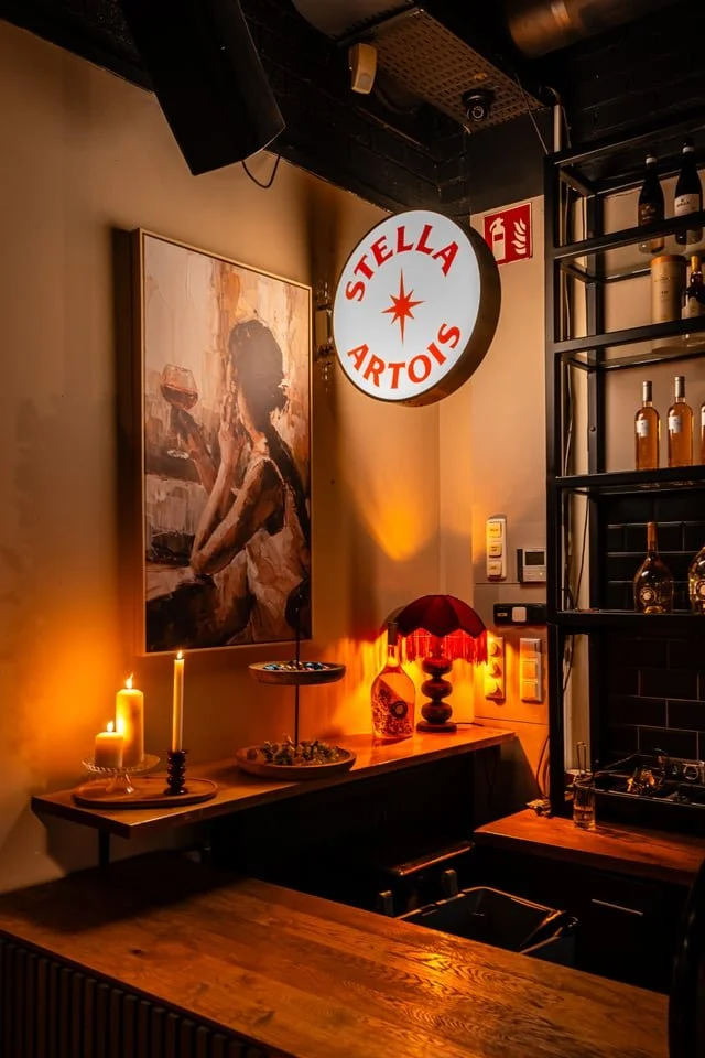 A cozy corner of a bar or restaurant with warm lighting, candles, and decorative items including a Stella Artois illuminated sign, a painting of a woman, a red lampshade on a table lamp, and bottles on a shelf.