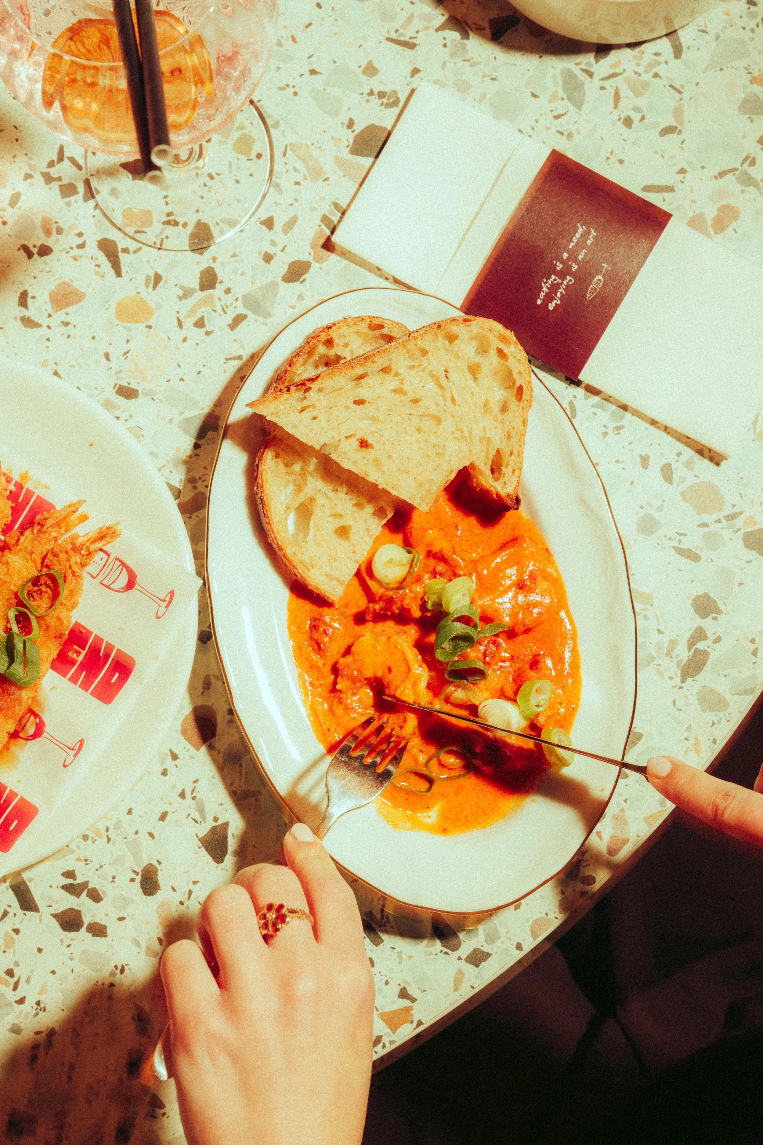 Plate of shrimp in orange sauce garnished with green onions, served with slices of bread, on a terrazzo table. A hand with a ring is reaching for the food, and another hand with a fork is about to take a bite.