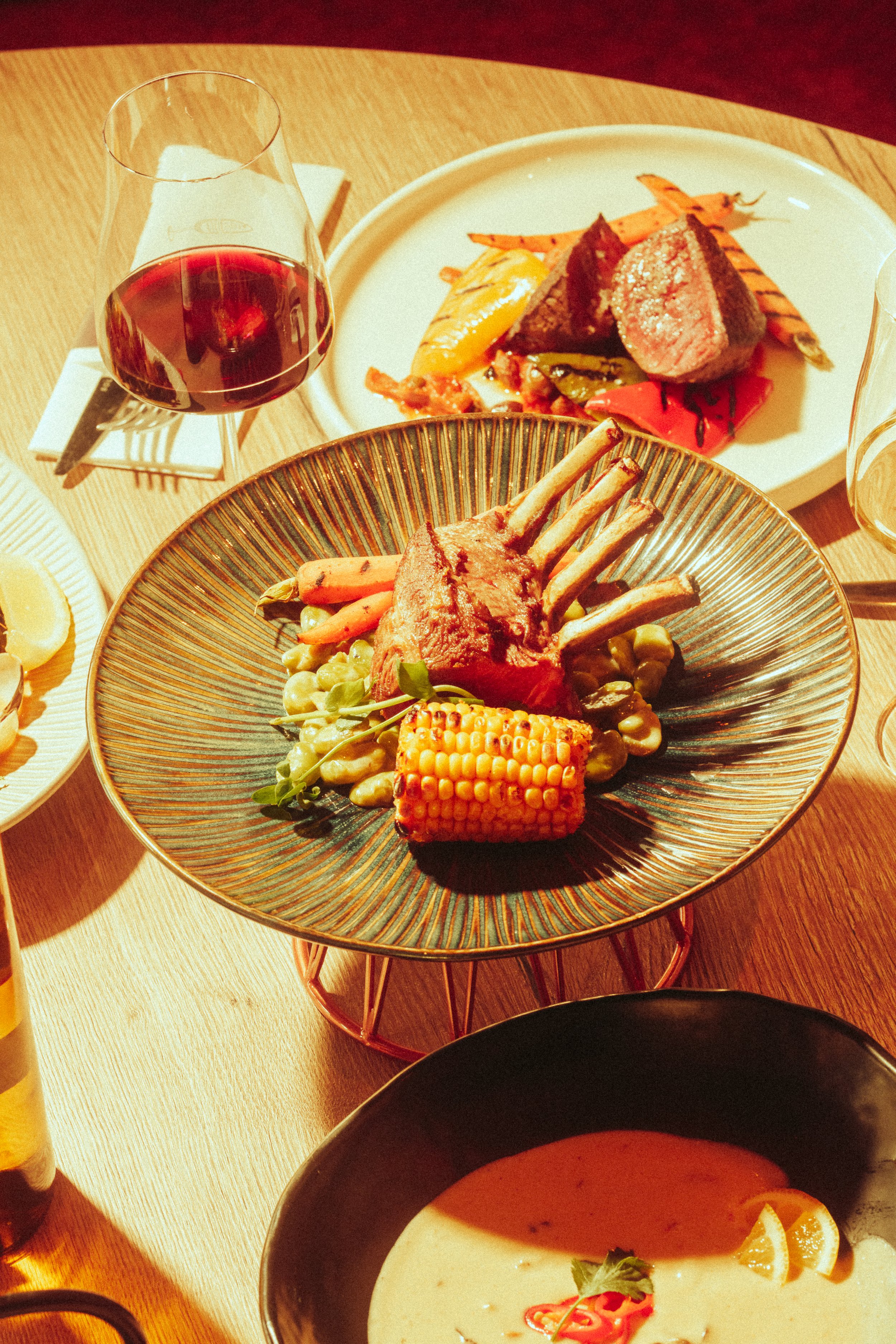 A plate of roasted lamb chops, corn on the cob, and vegetables on a table with a glass of red wine, a bowl of soup, and another plate of steak and grilled vegetables.
