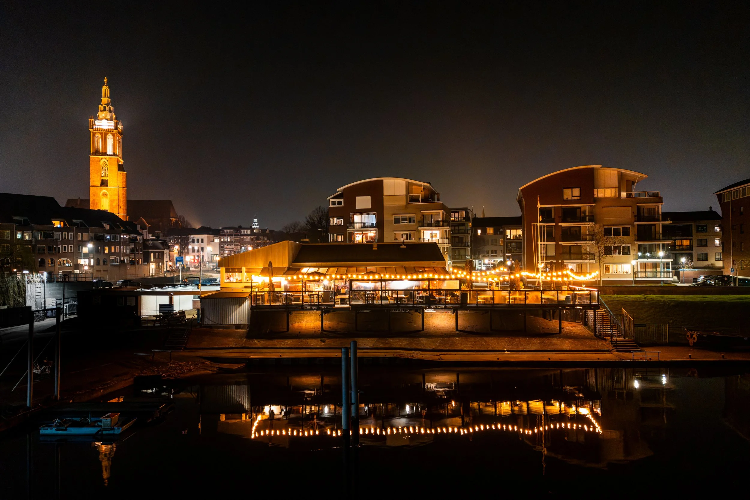 Night view of a waterfront area with illuminated restaurant or cafe, modern apartment buildings, and a historic church tower in the background, with reflections on the water.