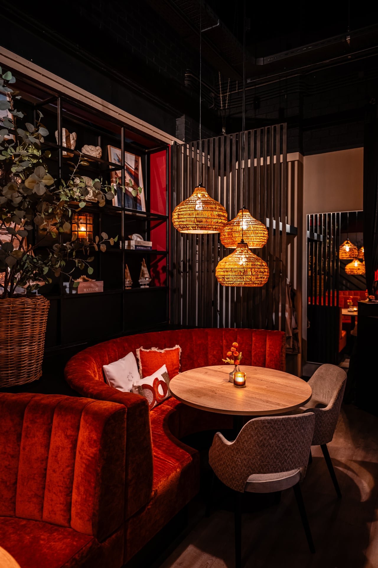 Cozy restaurant corner with a red velvet curved banquette, a round wooden table, and three hanging wicker pendant lights. Decor includes pillows on the banquette, a small flower arrangement, and a bookshelf with framed photos, books, and decorative i