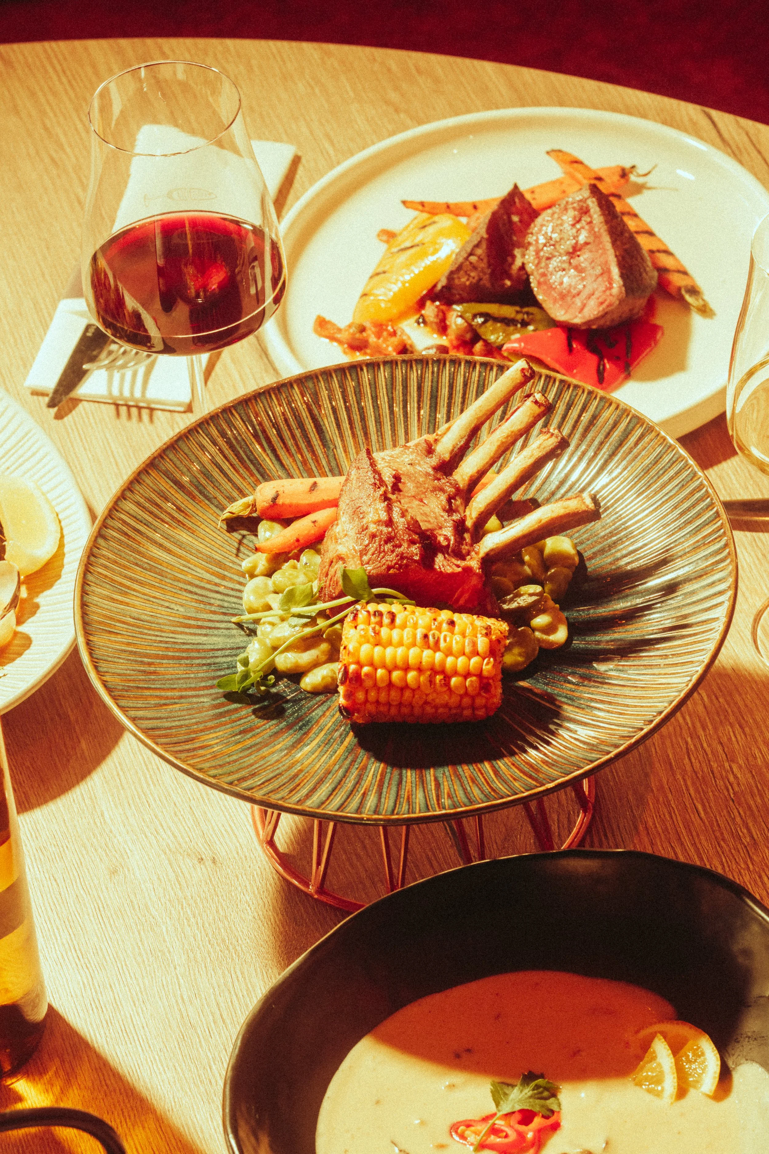 A plated meal with a main dish of lamb chops, vegetables, and corn, accompanied by a glass of red wine and other dishes in the background.