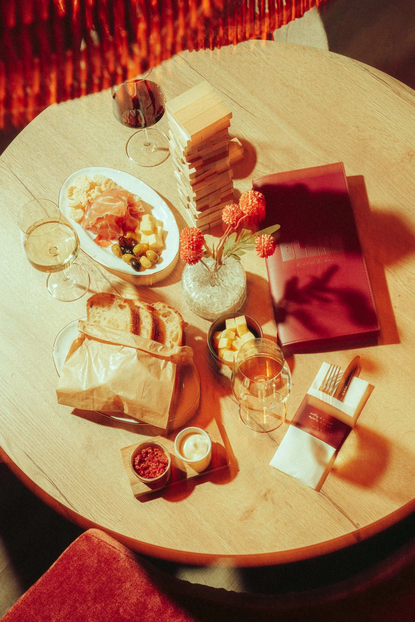 A round wooden table set for a meal, with a cheese platter, sliced bread, glasses of white wine, a small bowl of butter, a jar of red condiment, and a small dish of spread. There are decorative orange flowers in a glass vase, a stack of small cream-c