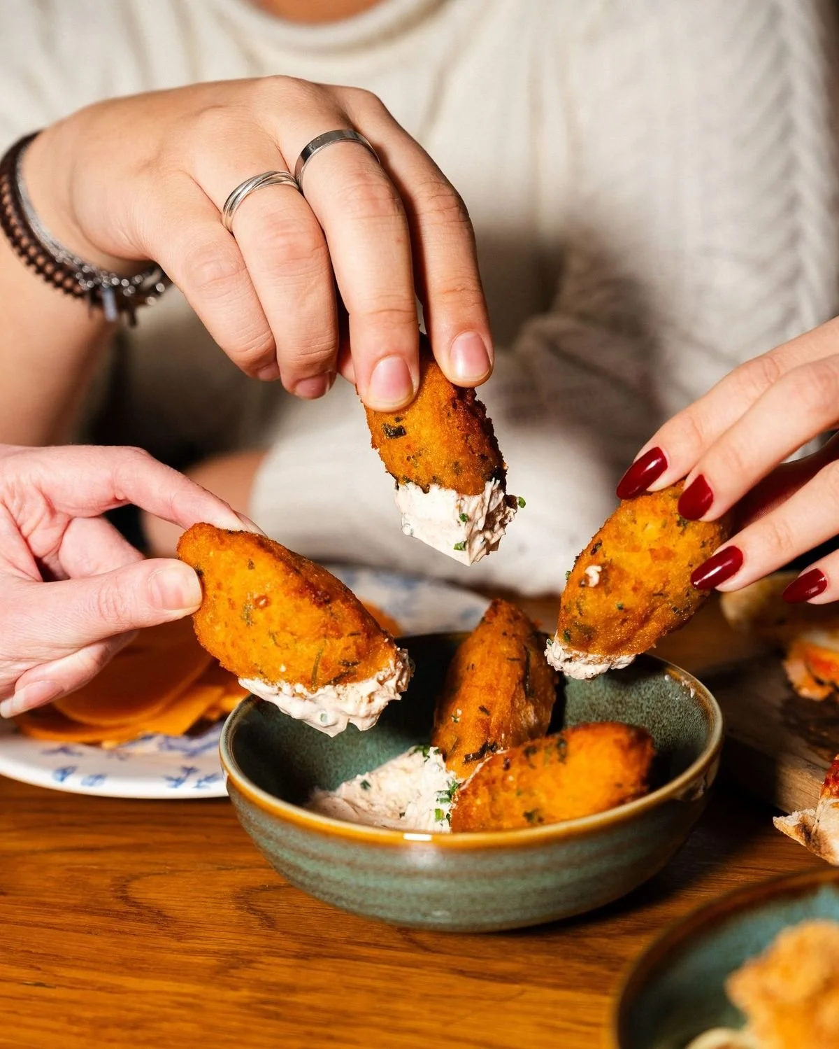 Two people dipping breaded, fried jalapeno poppers into a creamy sauce, with one person wearing multiple rings and bracelets and the other with painted red nails.