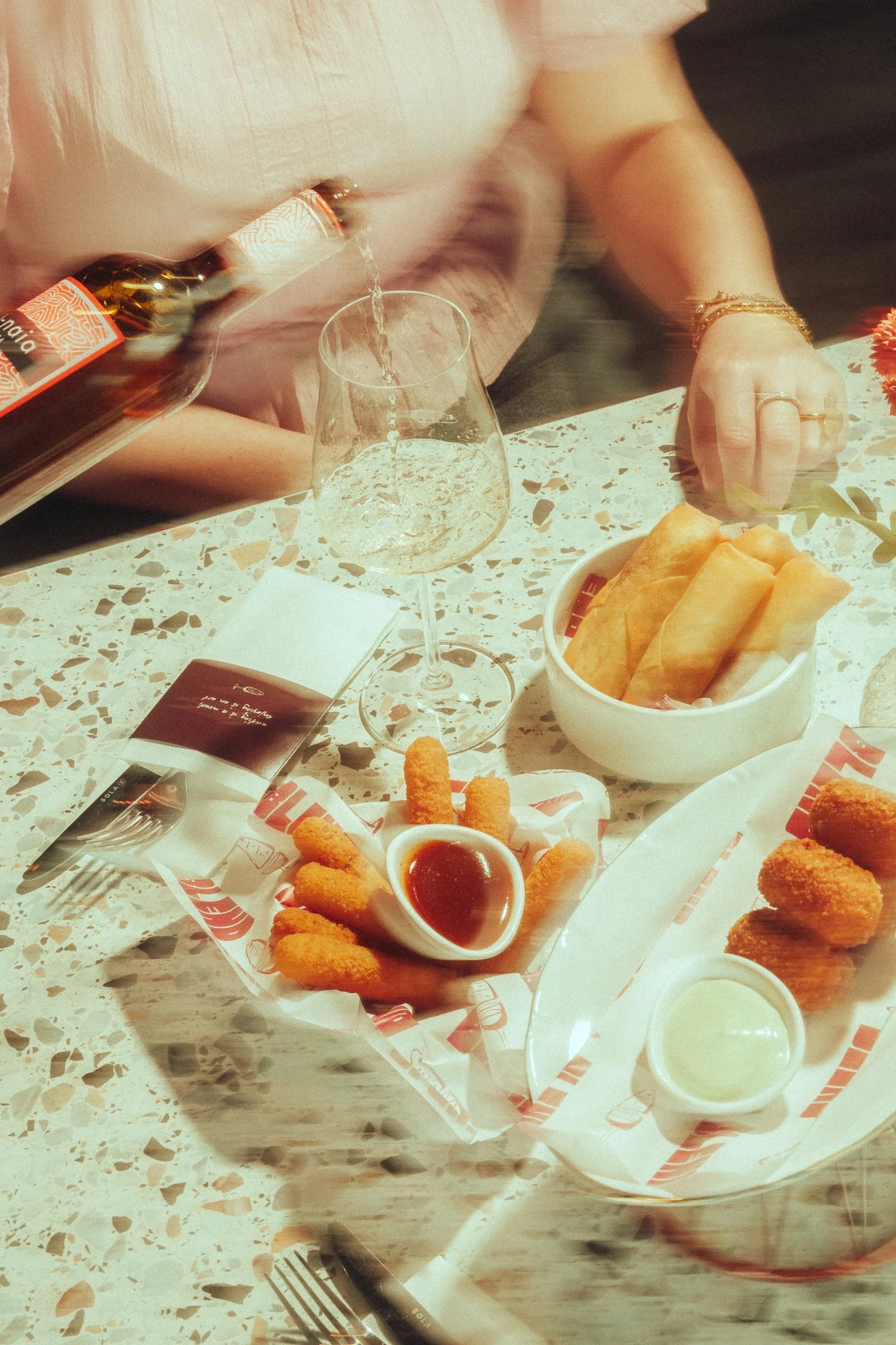 Person pouring a drink into a glass at a table with fried appetizers, spring rolls, and dipping sauces.