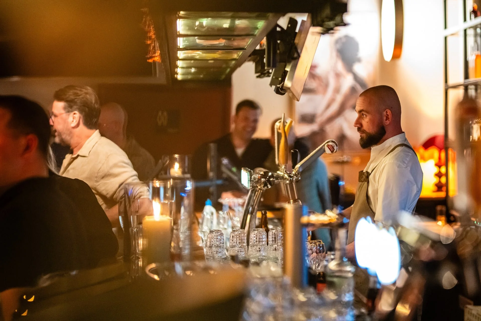 Barman with a beard and white shirt behind a bar counter, with several glasses and beer taps, in a warm, dimly lit bar with patrons in the background.