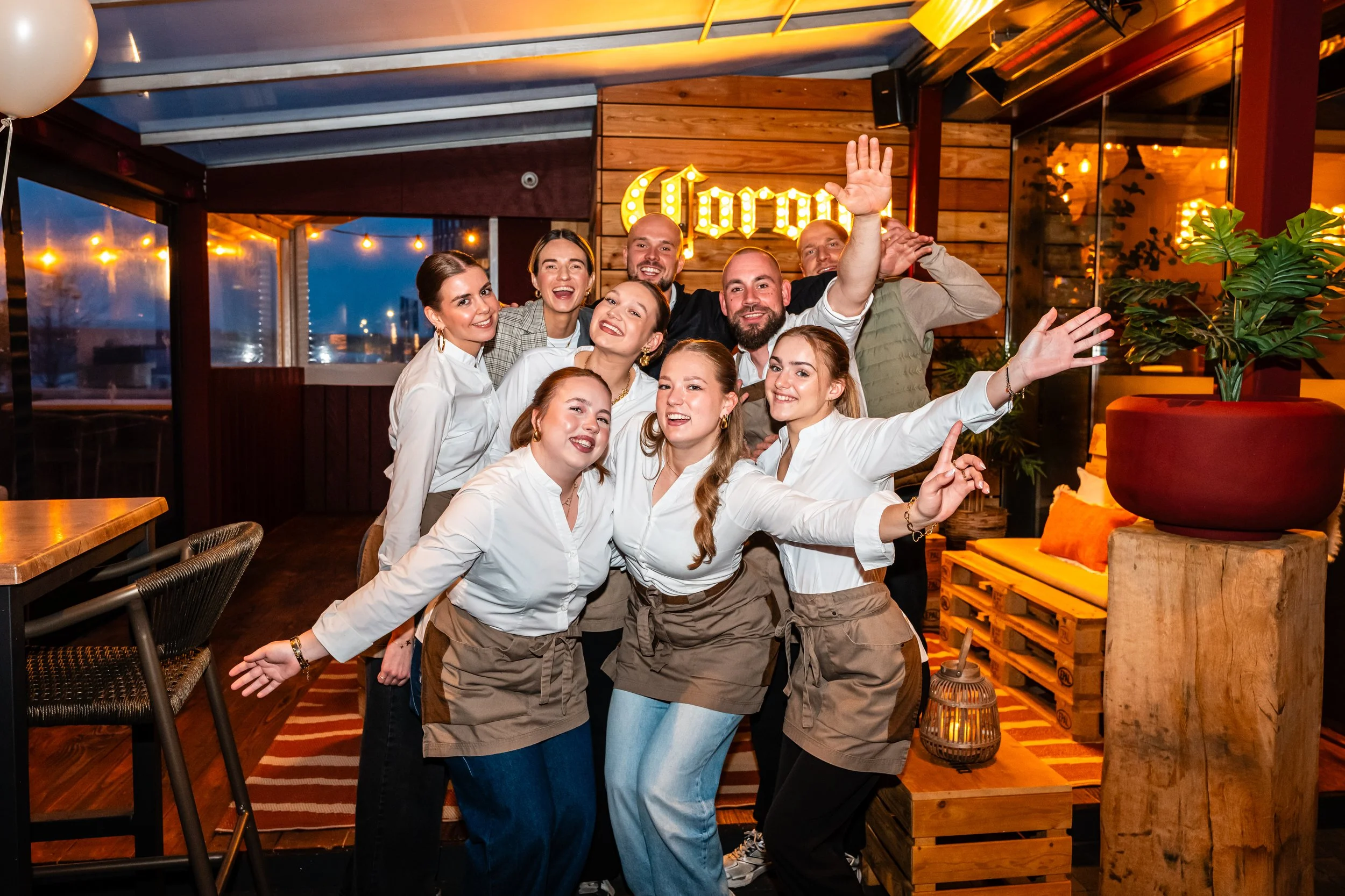 Group of happy restaurant staff posing together inside a warmly lit dining area, with a neon sign that reads 'Corona' in the background.