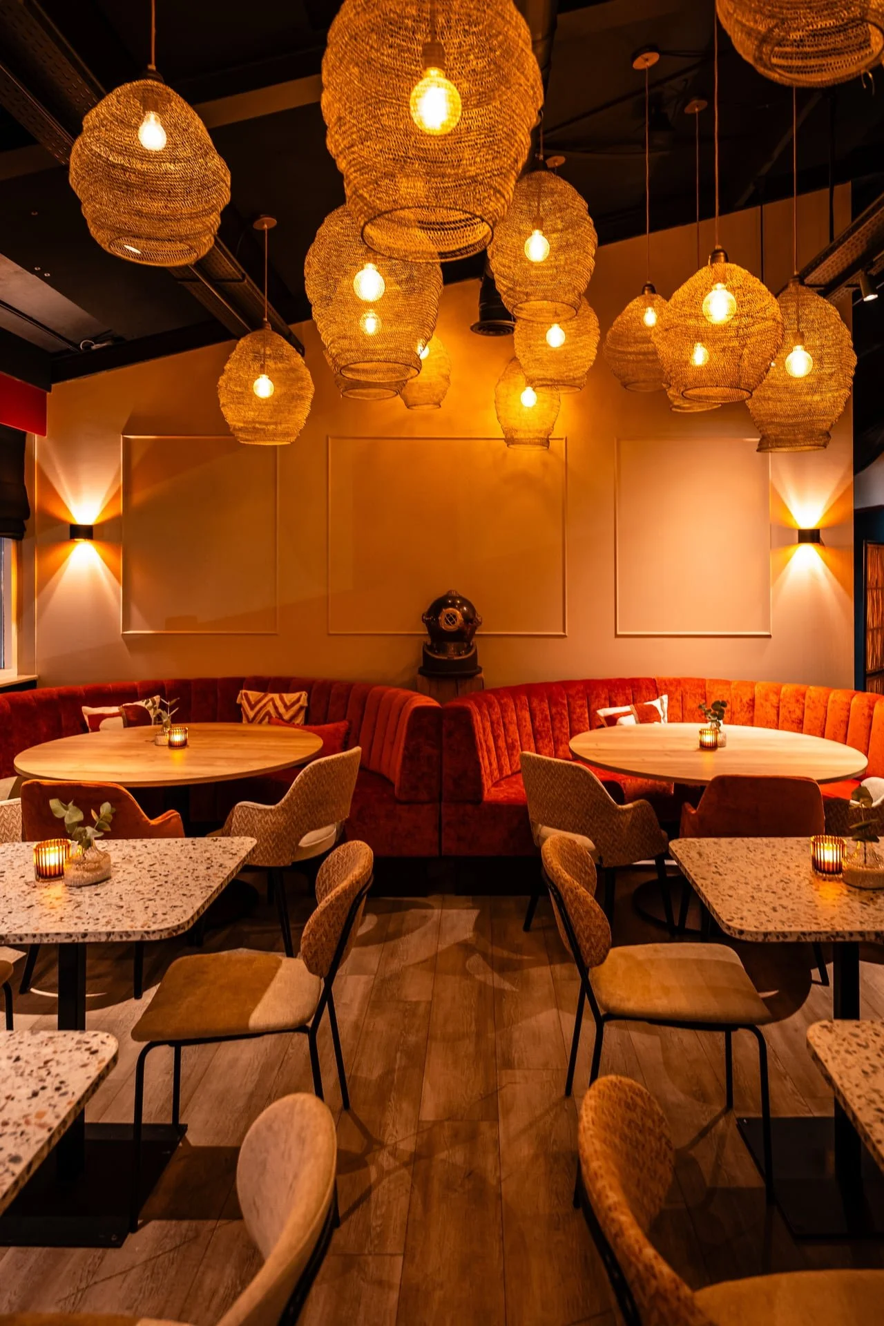 A cozy restaurant interior with warm lighting, hanging woven lamps, and a mix of dining tables, chairs, and a curved red velvet booth. Small candles and plants decorate each table.