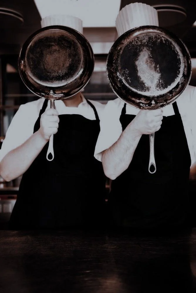 Two chefs wearing white uniforms, aprons, and chef hats holding skillets to their faces, standing in a kitchen.