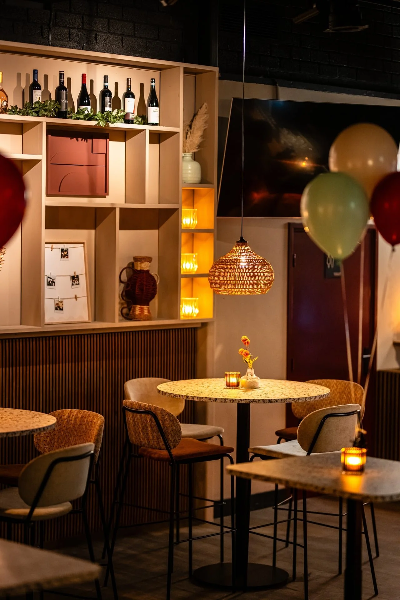 Cozy restaurant interior with round tables, chairs, and warm candle lighting, with a wall featuring decorative shelves, bottles, and a flat-screen TV.