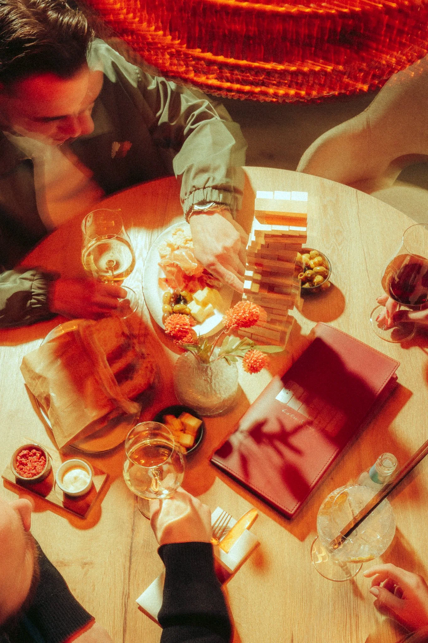 A group of friends gathered around a dining table playing Jenga, with drinks and snacks on the table, including cheese, grapes, and flowers.