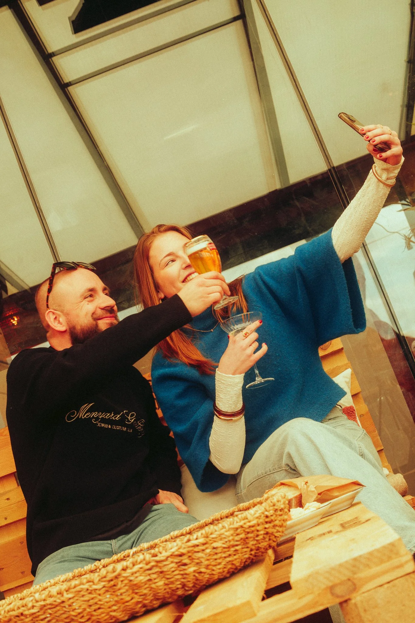 A man and woman sitting together on a wooden bench, taking a selfie while holding drinks, smiling and enjoying themselves indoors.