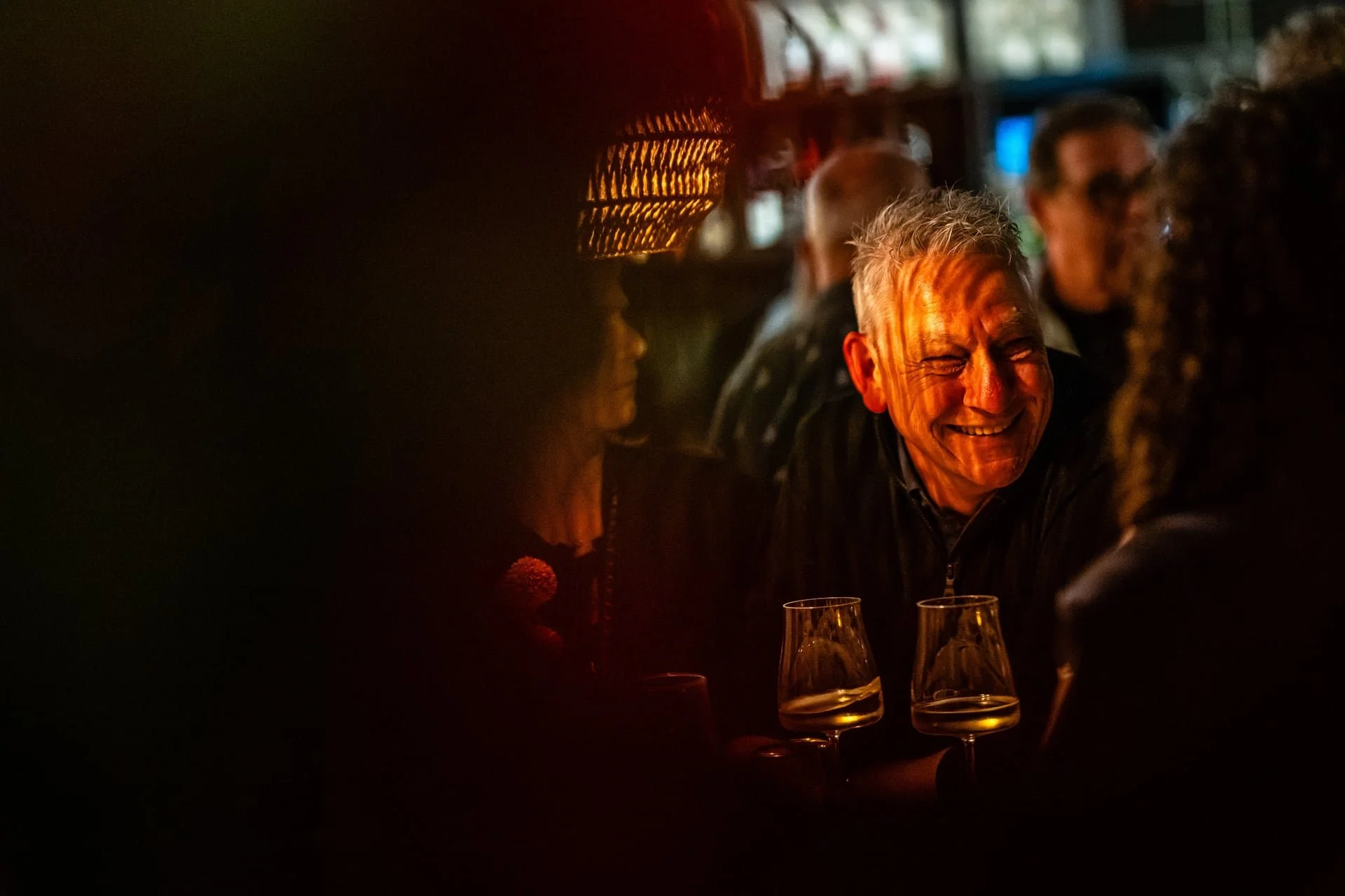 A group of people socializing in a dimly lit bar or restaurant, with a smiling older man in focus, enjoying conversation with others. There are wine glasses on the table.