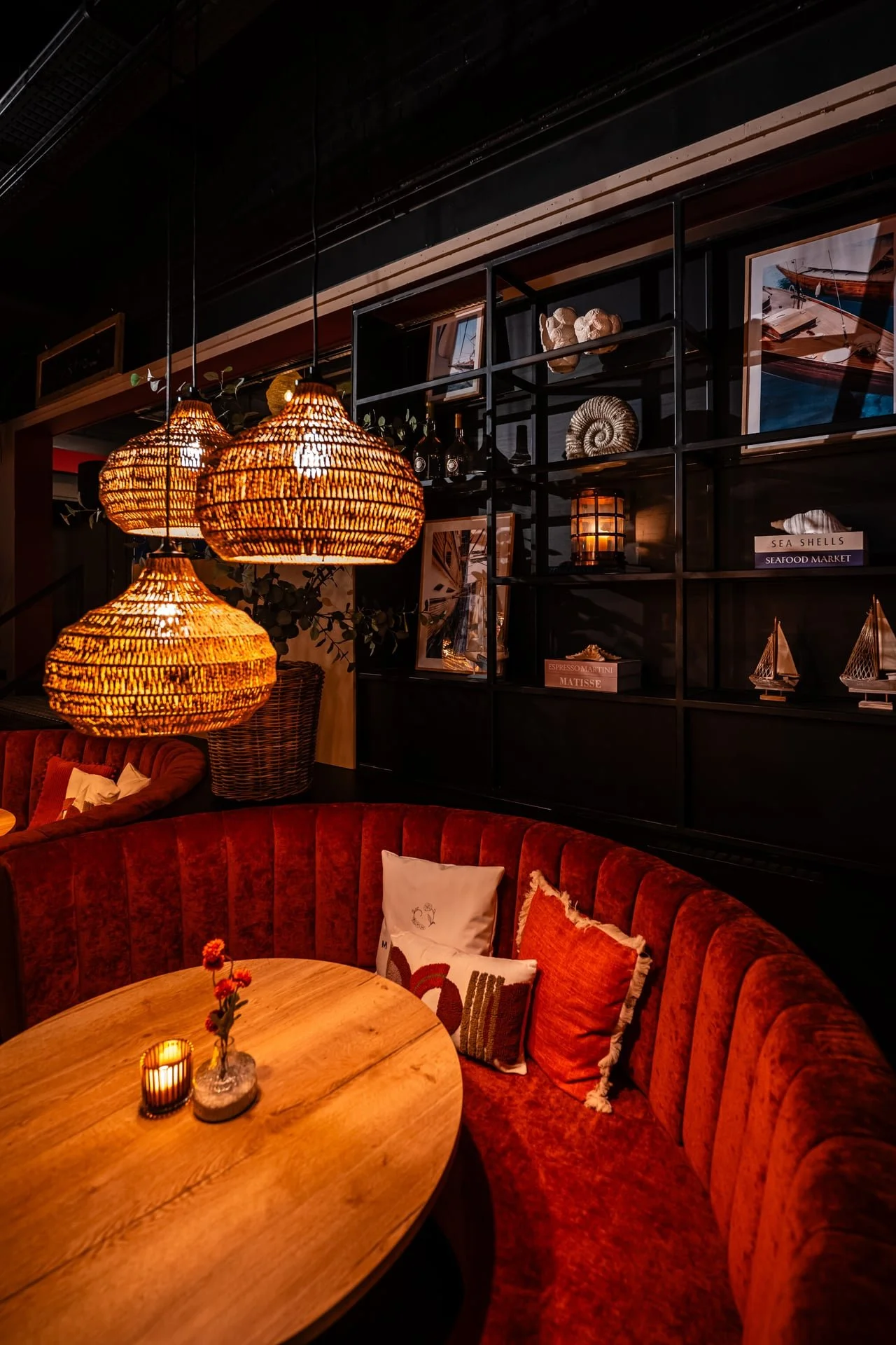 A cozy restaurant corner with a curved plush red banquette, wooden table with a candle and small flowers, and wicker pendant lights hanging from the ceiling. Behind are black shelving units with nautical-themed decorations and framed photos.